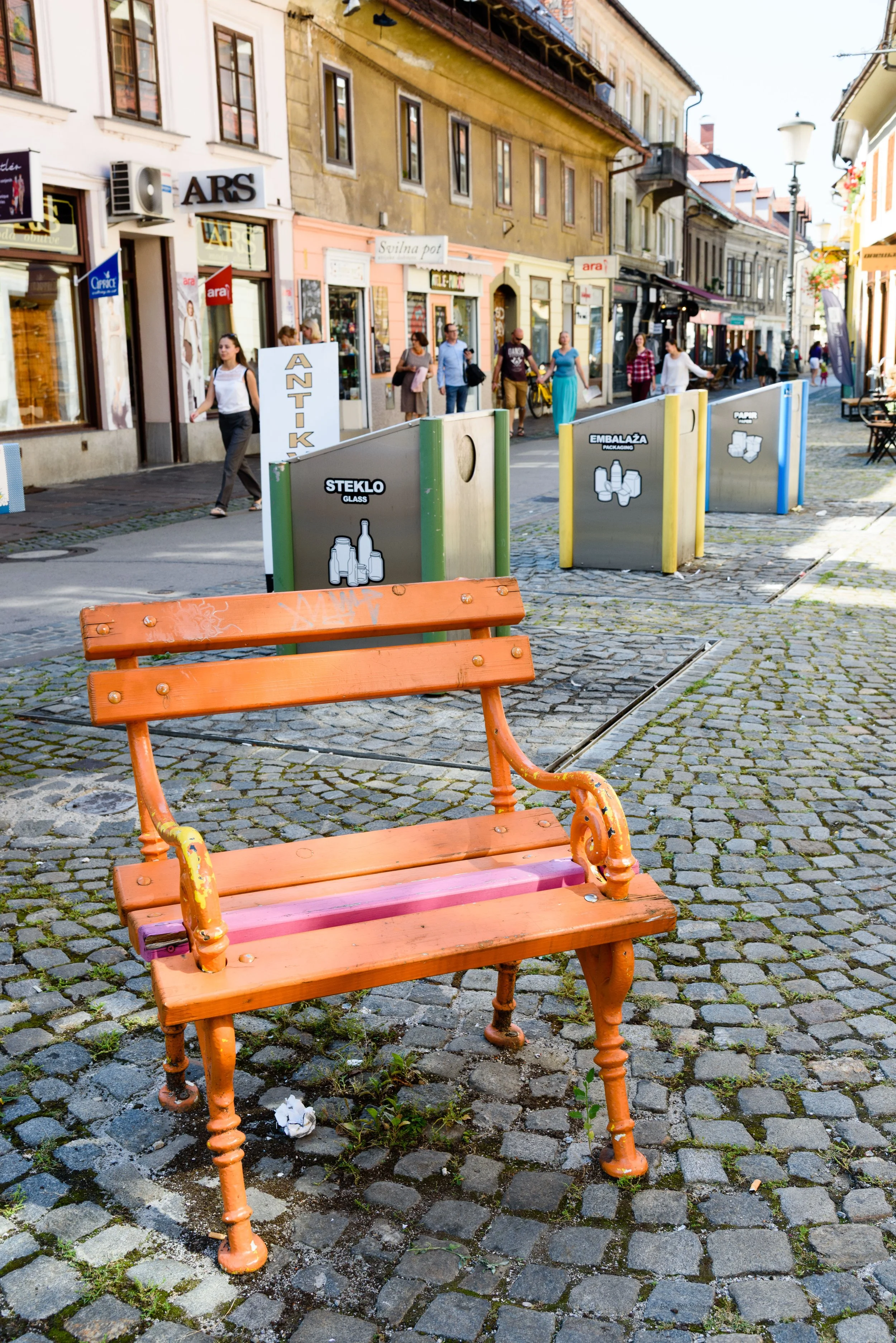 Empty orange park bench on cobblestone street in a city with people walking and shops in the background.
