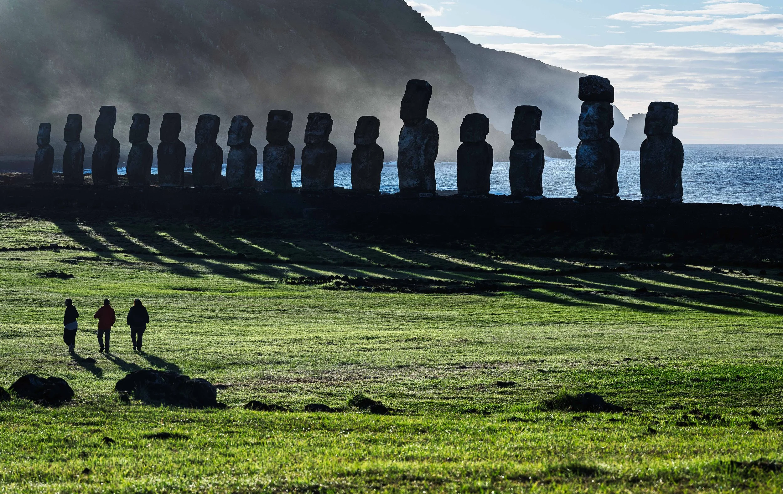 Three people walk across a grassy field toward a row of Moai statues on Easter Island, with misty hills and the ocean in the background.