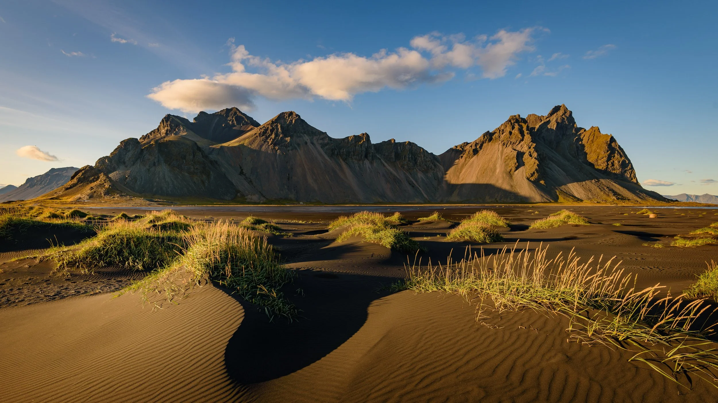 Mountains over a sandy desert landscape with patches of green grass and a clear blue sky.