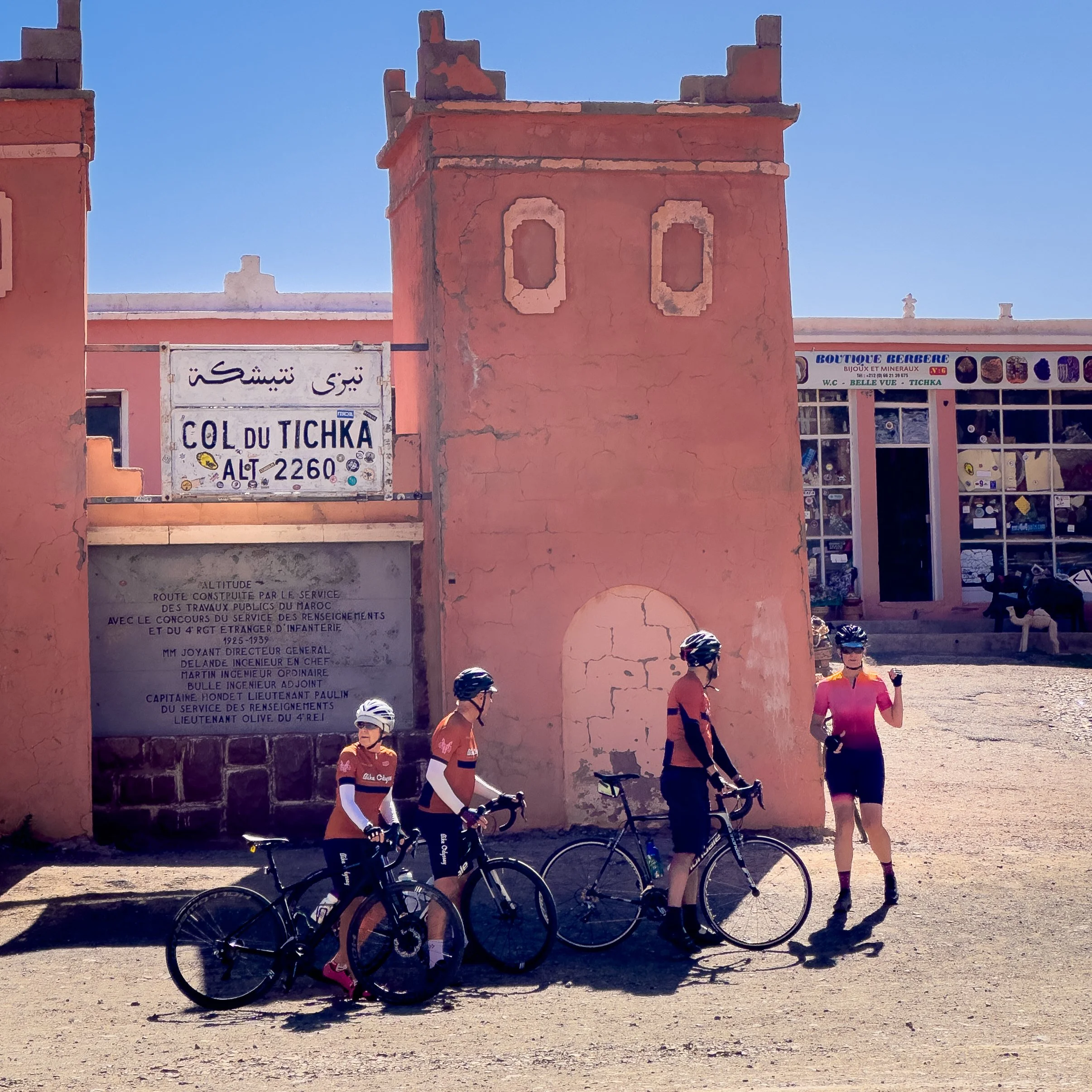 Four cyclists in matching orange and pink jerseys and helmets standing with their bikes in front of a pink building with signs that read "Col du Tichka" and "Boutique Berbere" in a sunny outdoor setting.