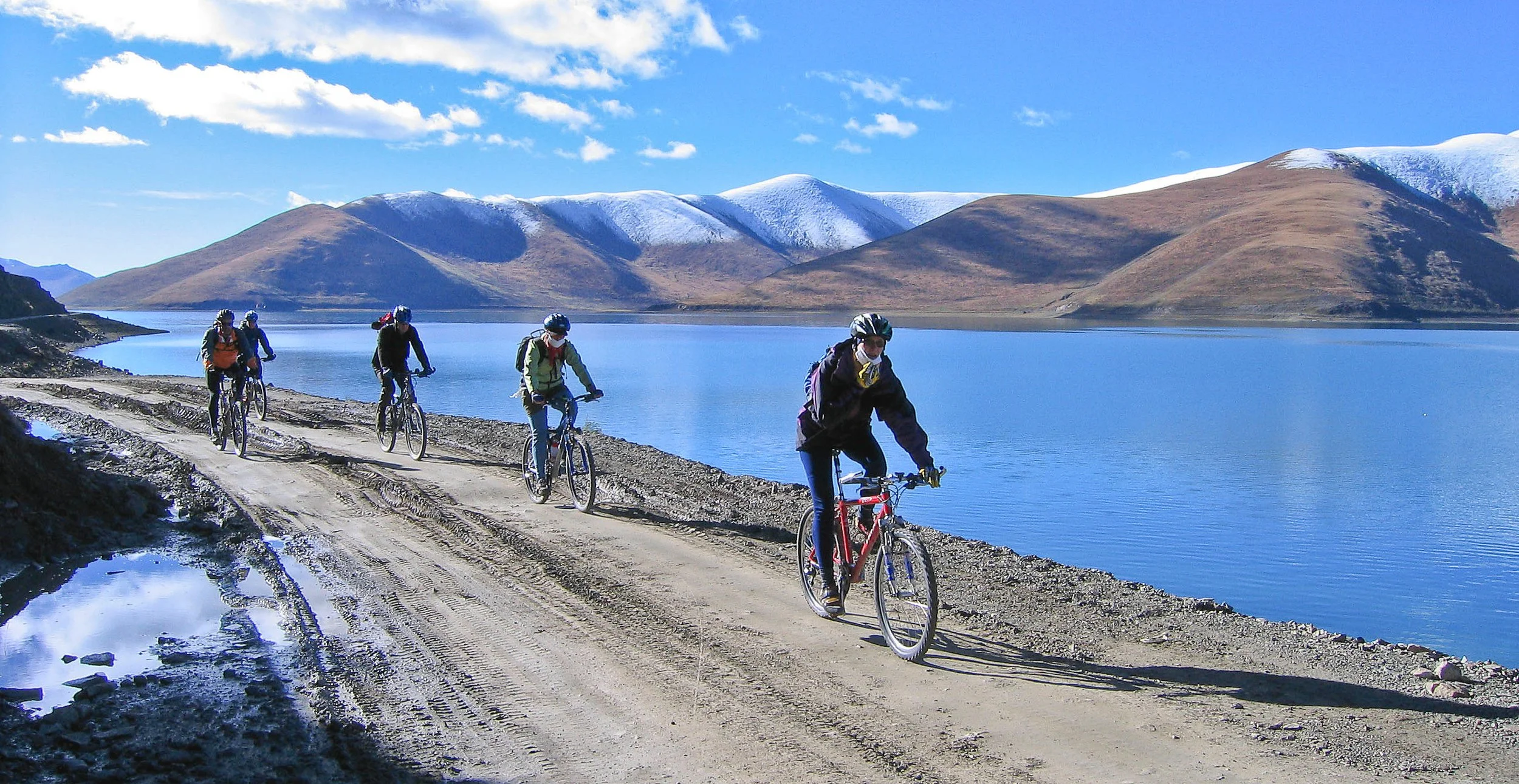 Four cyclists riding on a dirt road alongside a large blue lake with mountains in the background under a partly cloudy sky.