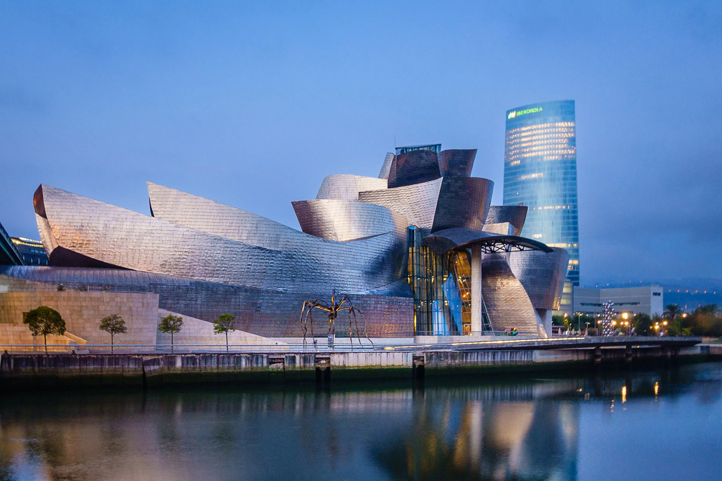 The Guggenheim Museum Bilbao with a reflective water body in the foreground, a spider sculpture nearby, and a tall office building with 'IBERDROLA' sign in the background, during twilight.