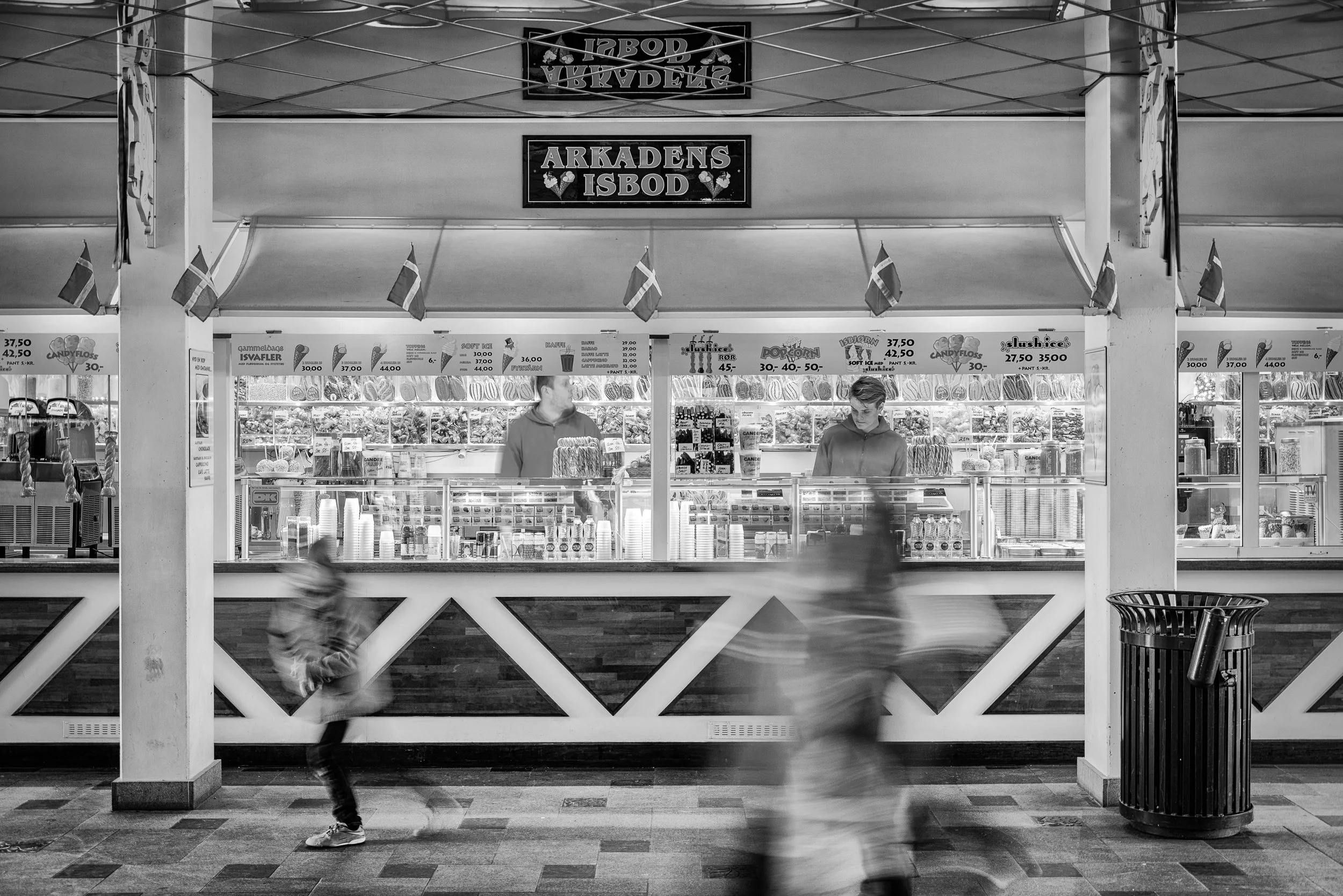 Black and white photo of a candy and snack kiosk in a shopping mall with two workers behind the counter and blurred children passing by in the foreground.