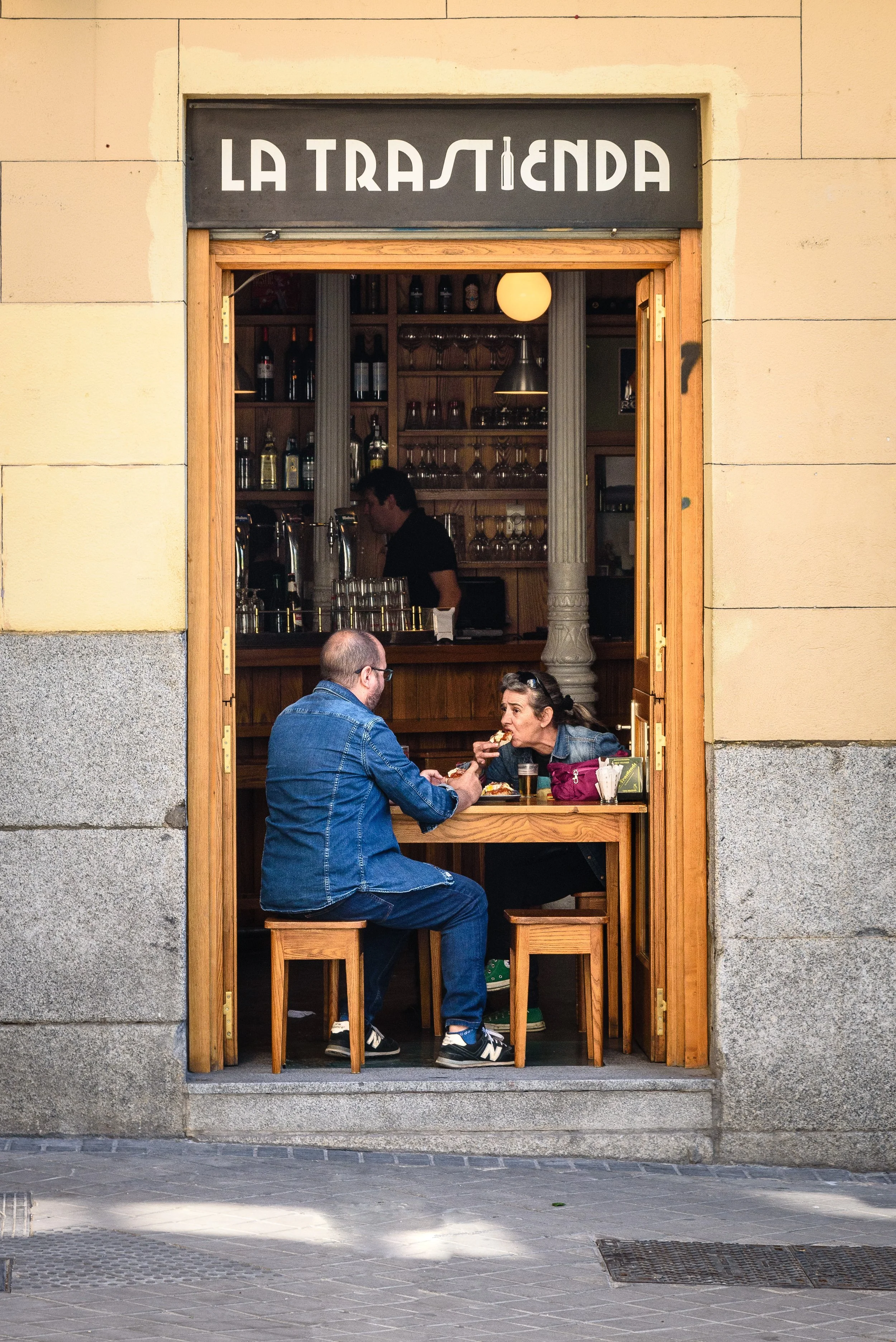 Two people are sitting at a small table inside a cozy restaurant named La Trastienda, sharing a meal and drinks. The restaurant has a wooden interior with shelves of bottles and glasses in the background, and an open doorway framed with wood leads to