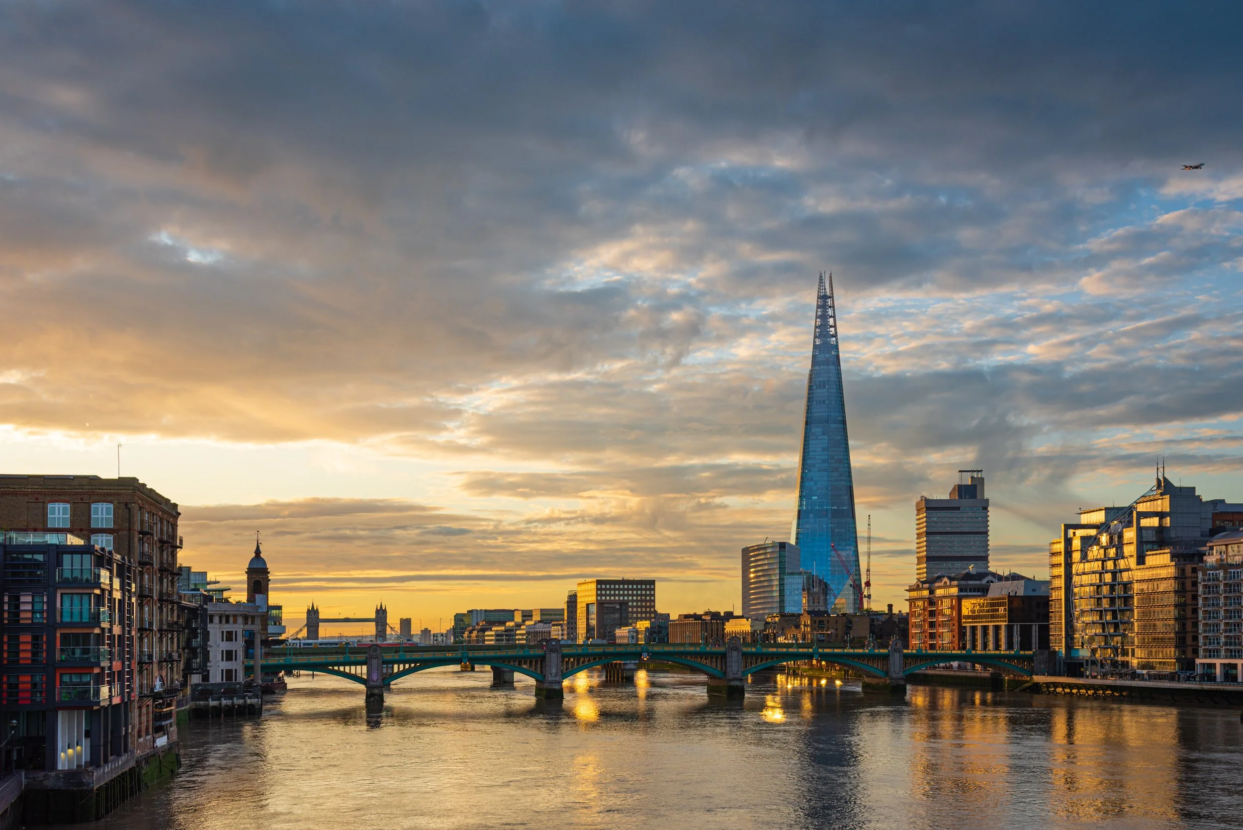 Sunset view of London skyline featuring the Shard skyscraper with river in foreground and bridge crossing it.