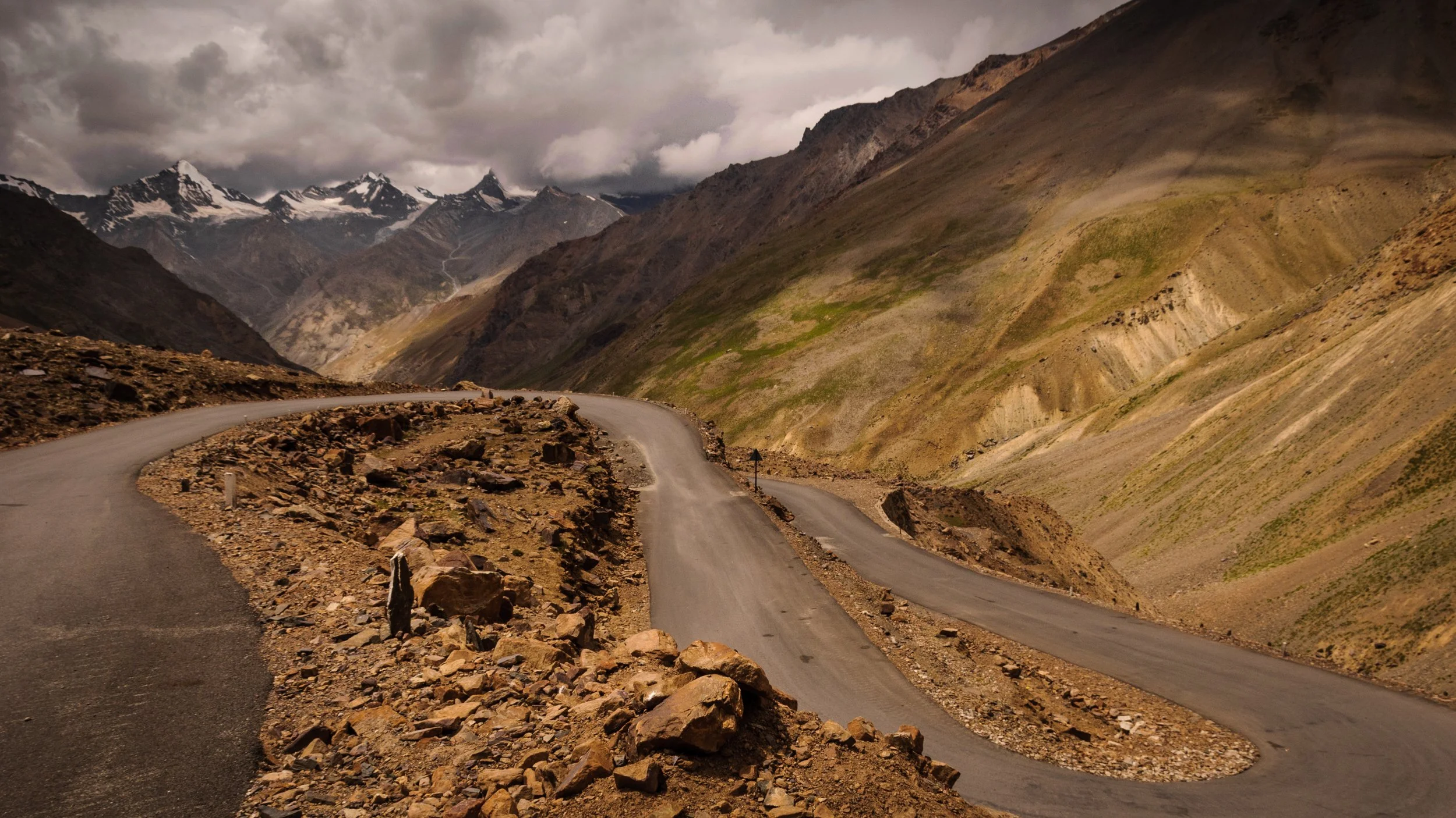 Winding mountain road with rocky terrain and green slopes, snow-capped peaks in the background, and dark cloudy sky.