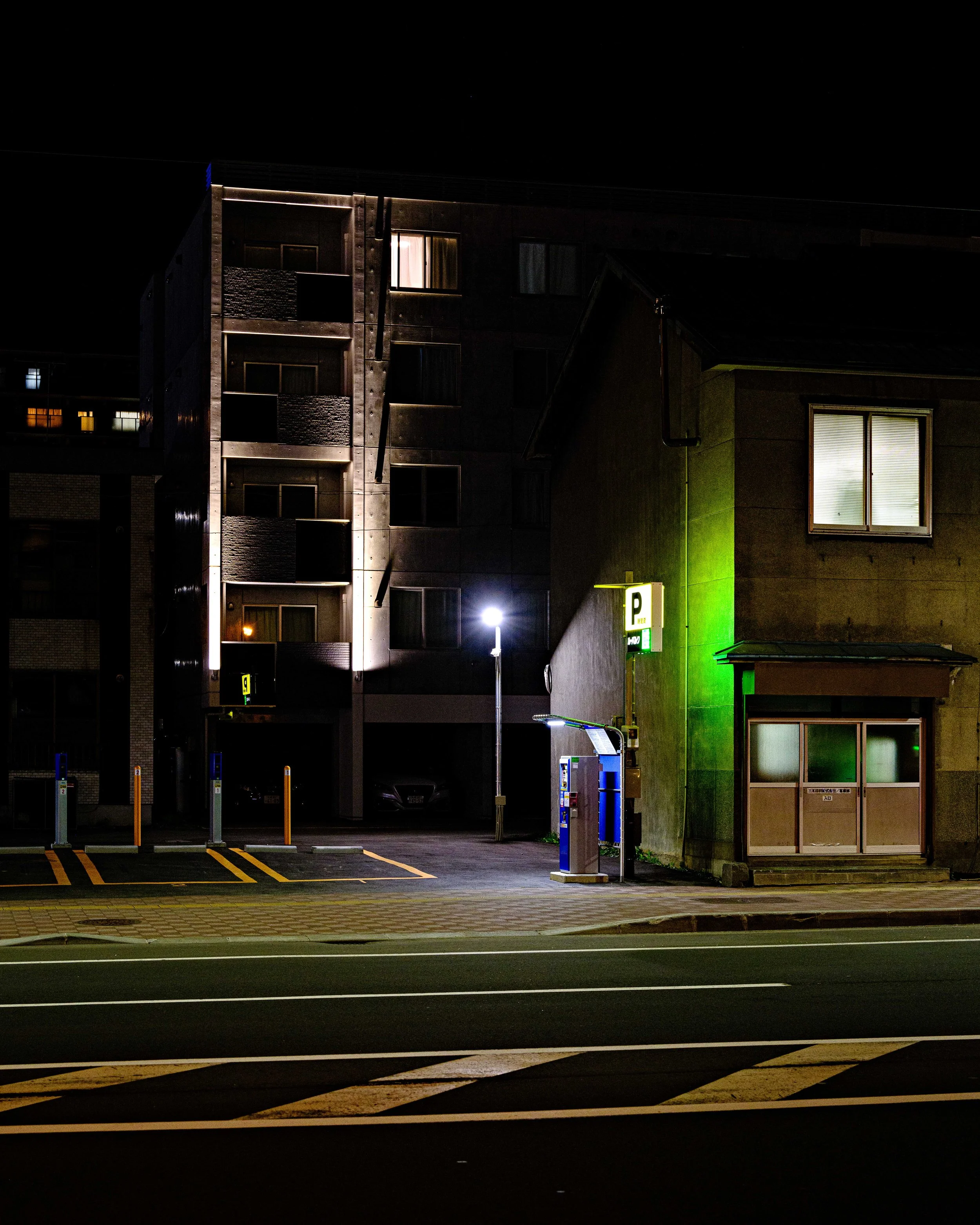 A quiet, well-lit parking lot at night with a parking meter, green neon sign, and surrounding buildings