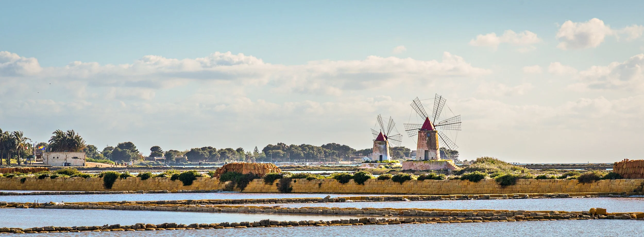 Salt ponds with two traditional windmills and a small white building in the background, under a partly cloudy sky.