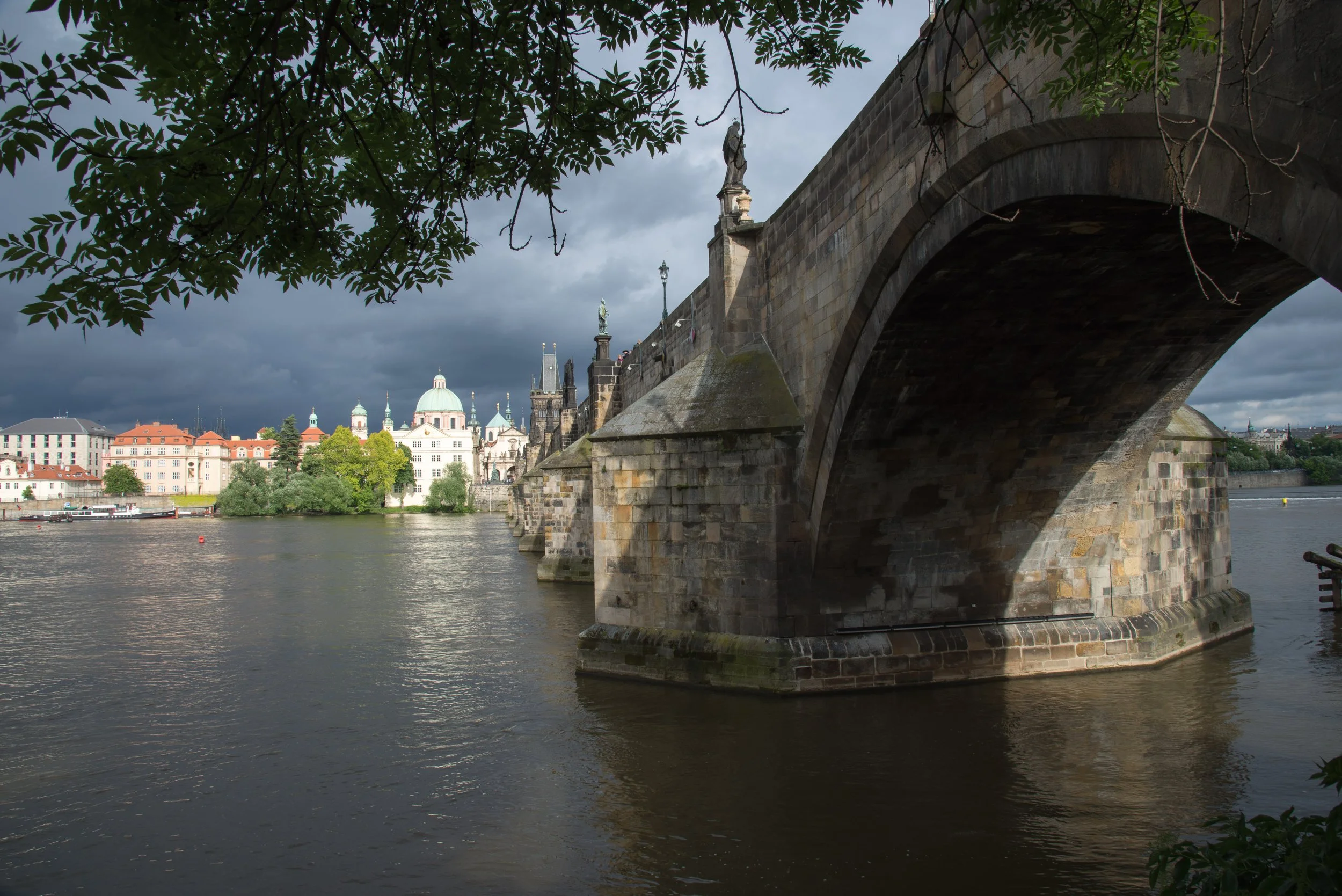 View of a stone bridge over a river in a city with historic buildings and a cloudy sky in the background.