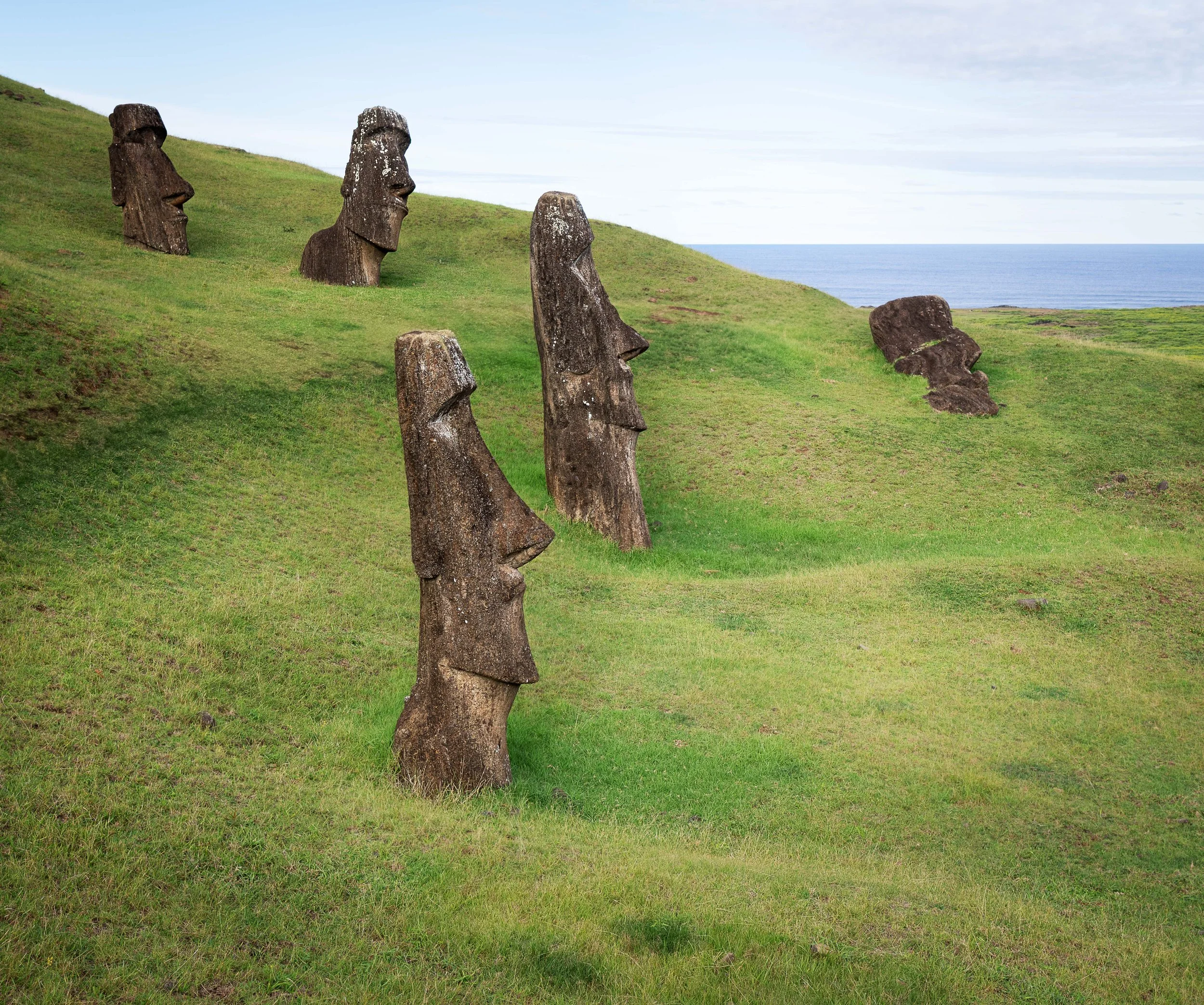 Five Moai statues on a grassy hillside with ocean in the background, hillside has varying green shades, and sky is partly cloudy.