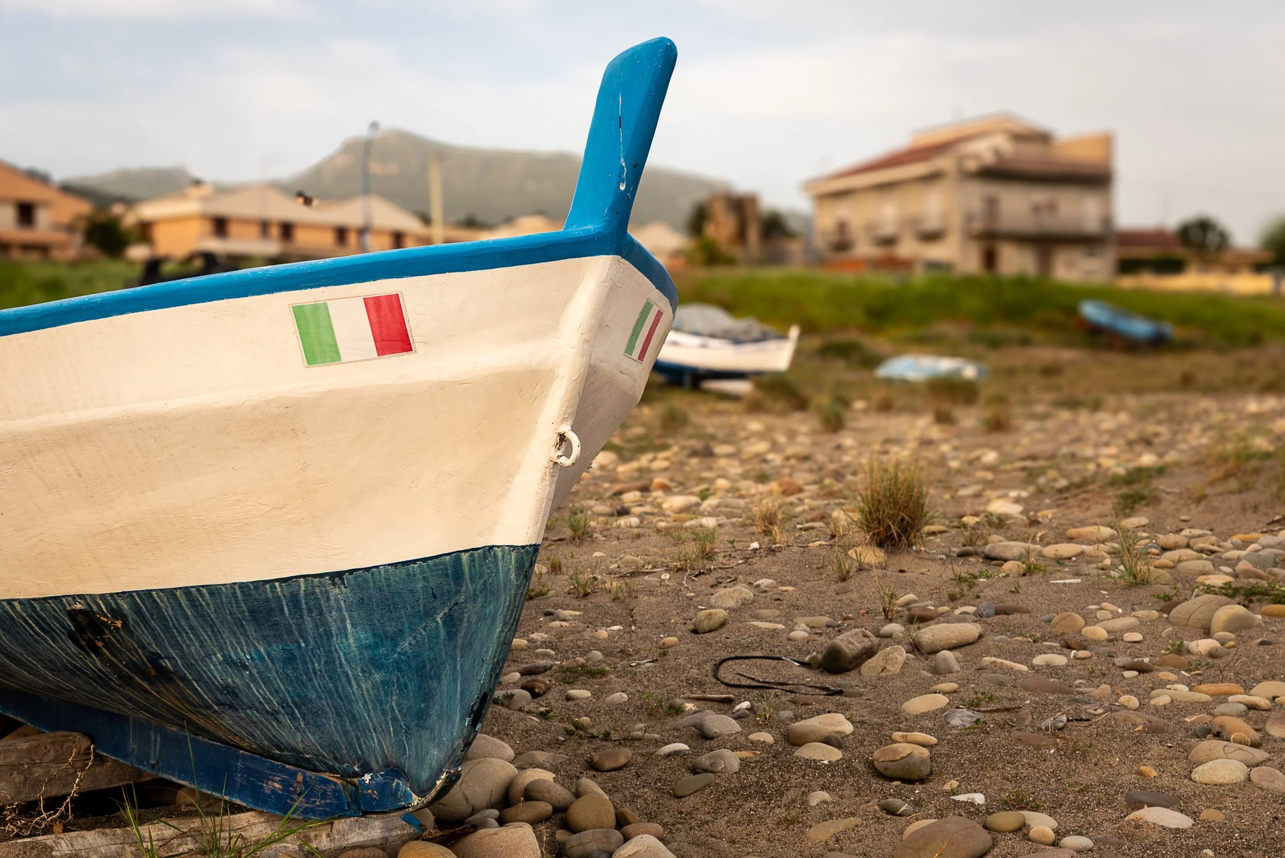Close-up of a small fishing boat with an Italian flag on the hull, resting on pebbly ground near houses in the background.