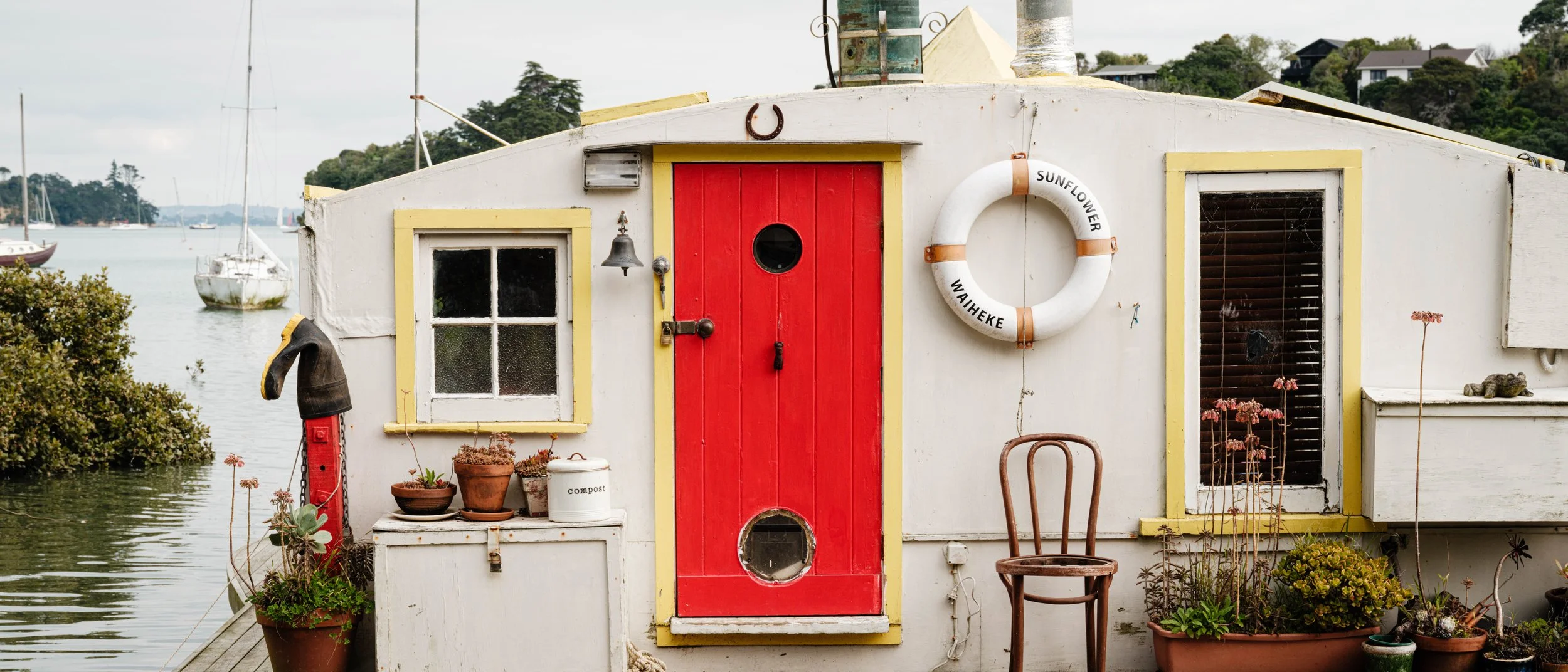 A white houseboat with yellow trim and a red door, decorated with potted plants, a life ring, and outdoor furniture, docked near sailboats on a calm body of water with sailboats and trees in the background.