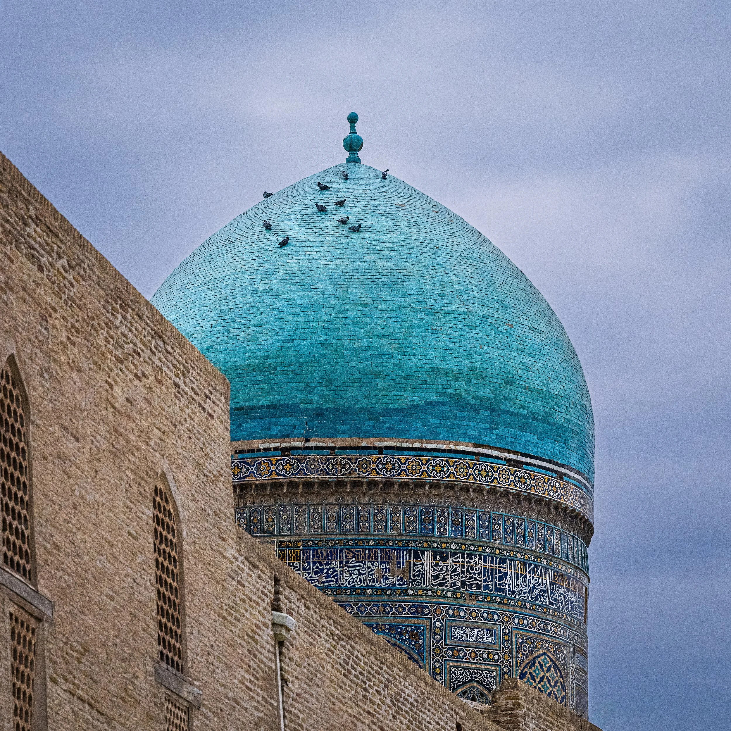 A blue-domed minaret with intricate tile work and Arabic script, partially obscured by a beige brick building, under a cloudy sky.