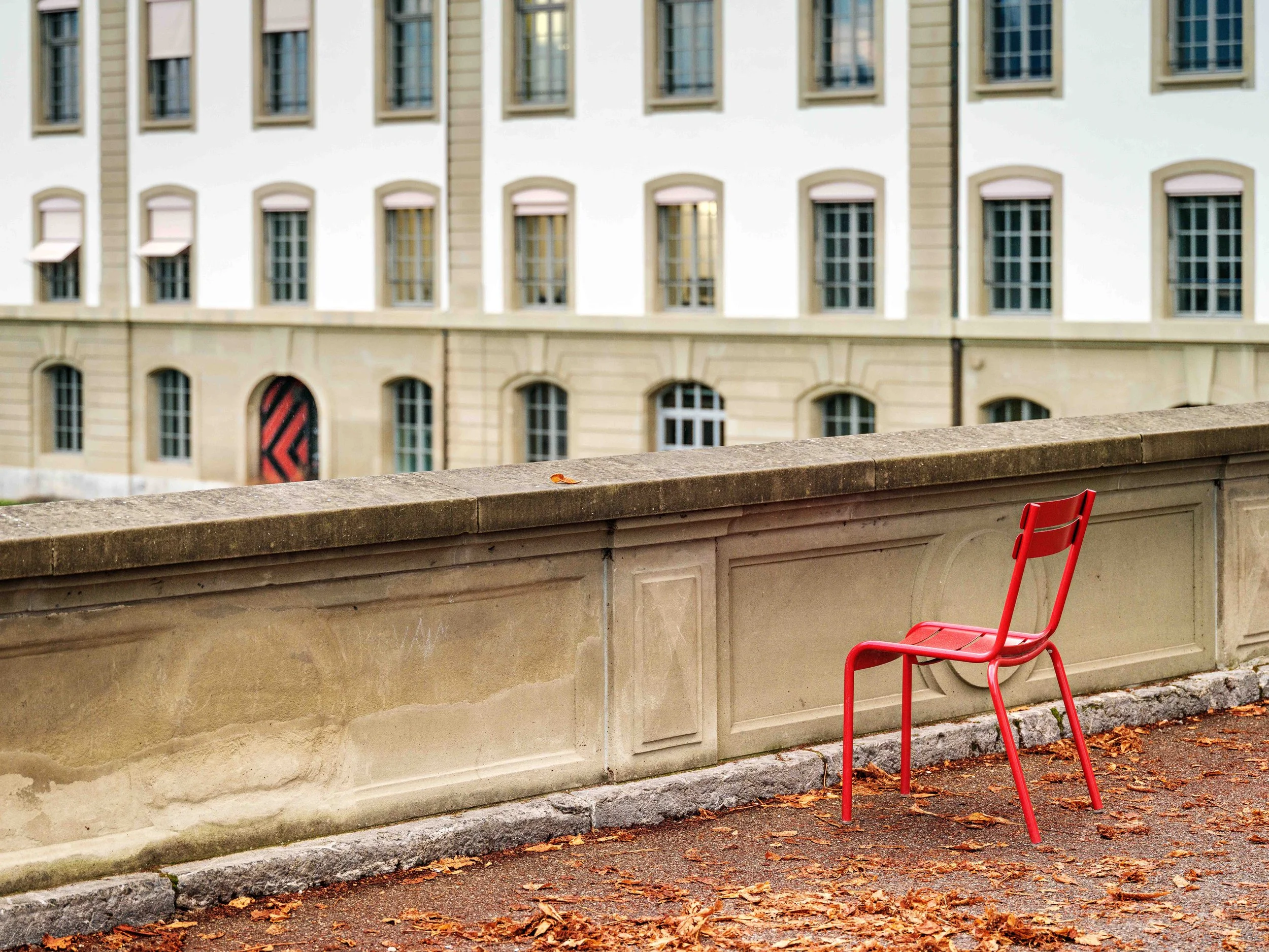 A red metal chair on a sidewalk with fallen autumn leaves, in front of a stone railing, overlooking a large beige and white building with multiple windows.