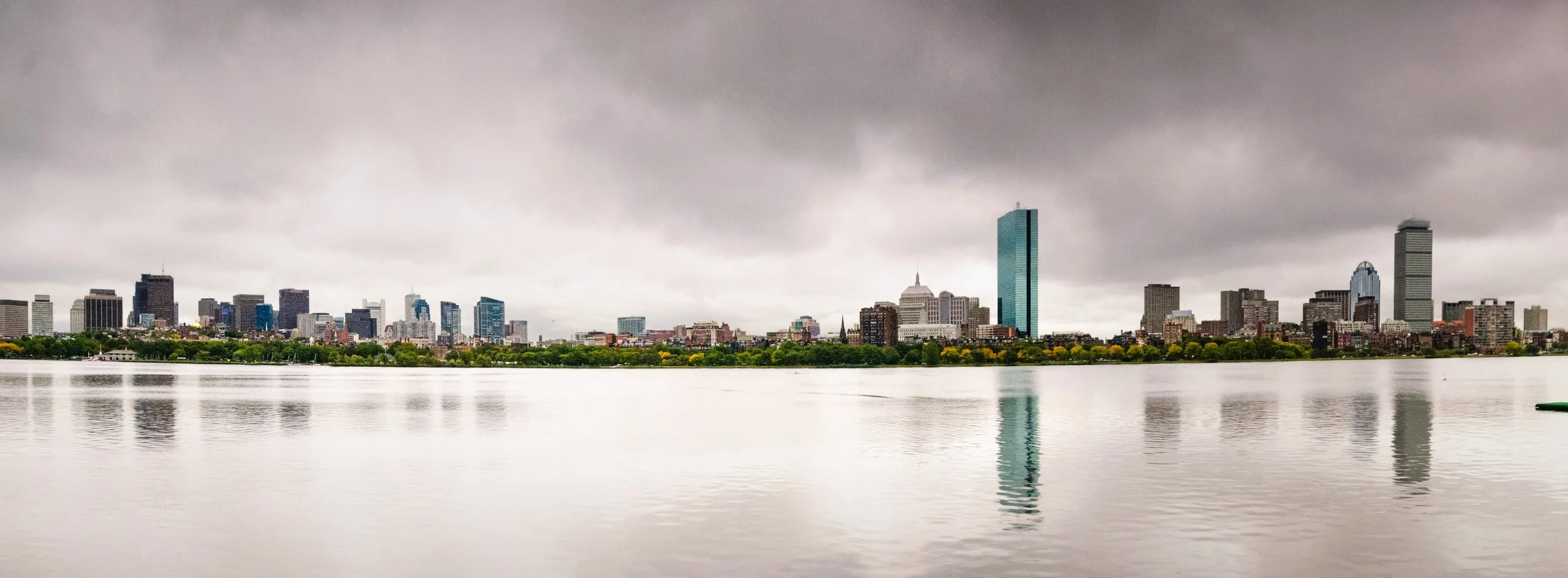 Cloudy skyline of Boston, Massachusetts, viewed across a body of water, featuring various skyscrapers including a tall, blue glass building.
