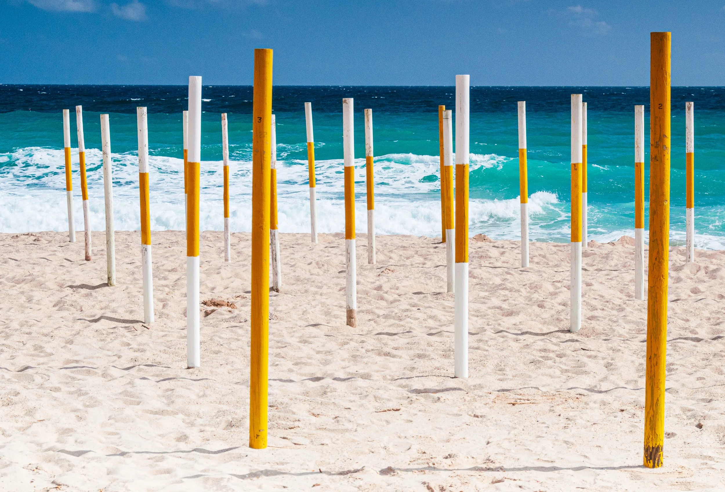 Beach with multiple white and yellow poles arranged in rows on sand, ocean waves in the background, blue sky overhead.