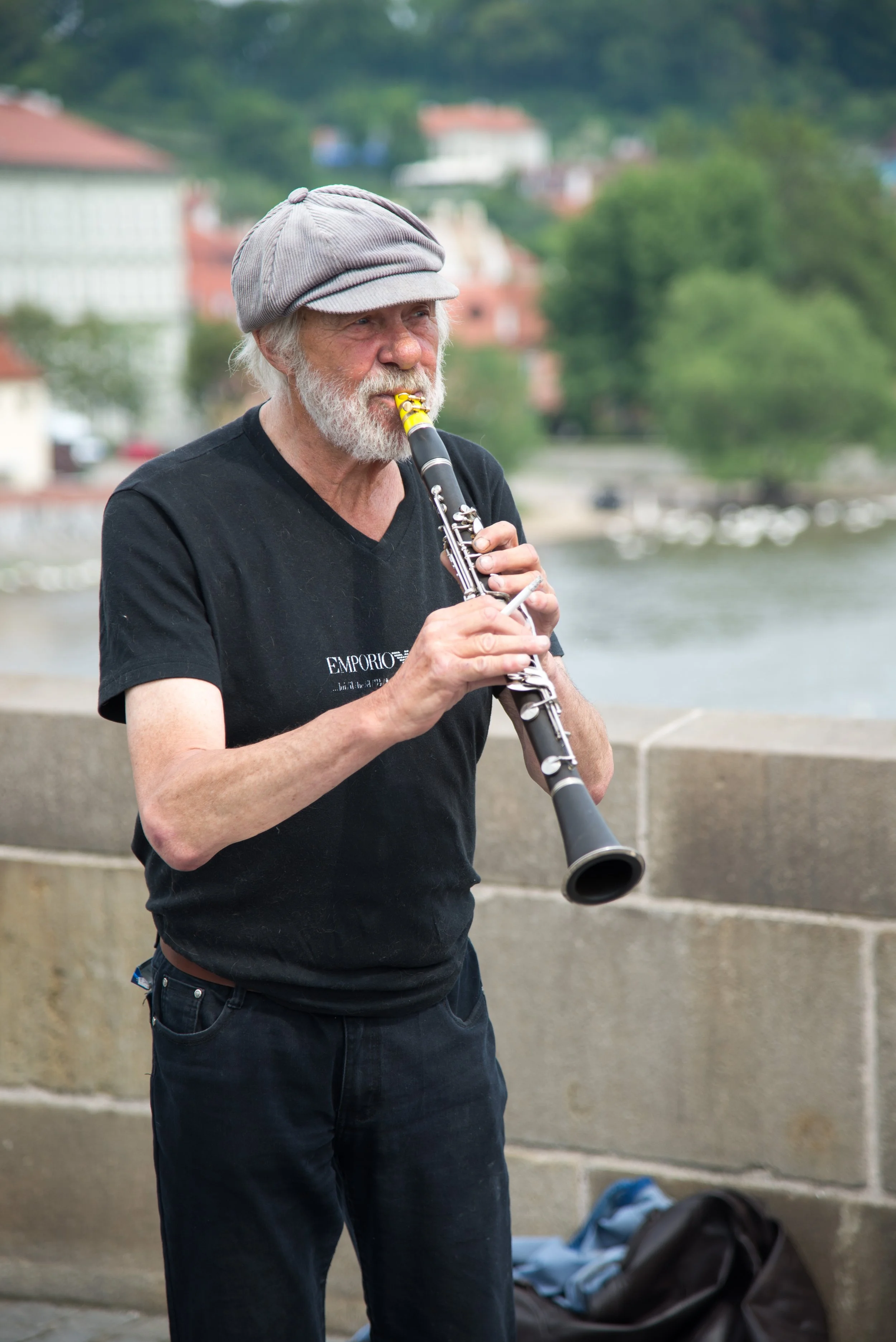 An elderly man with white hair and beard playing a clarinet outdoors near a river with buildings and trees in the background.