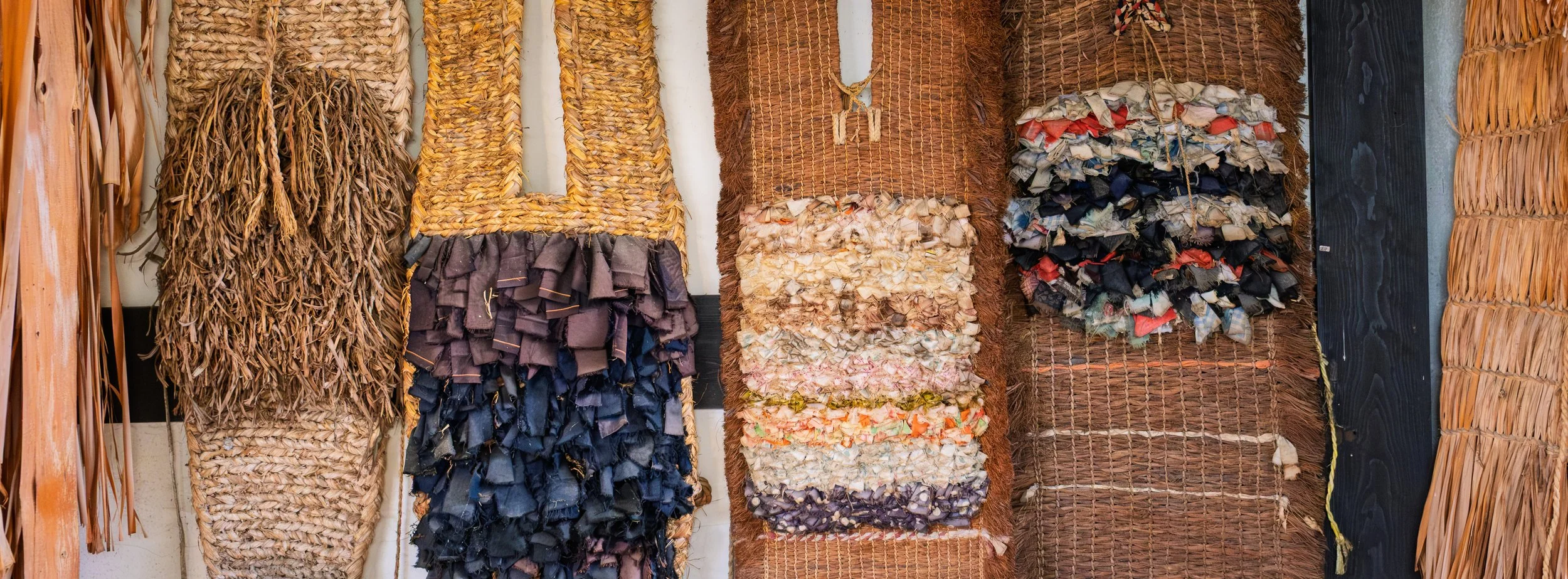 Rows of woven rugs or mats made from natural fibers, some with fabric scraps or strips attached, hanging on a wall.
