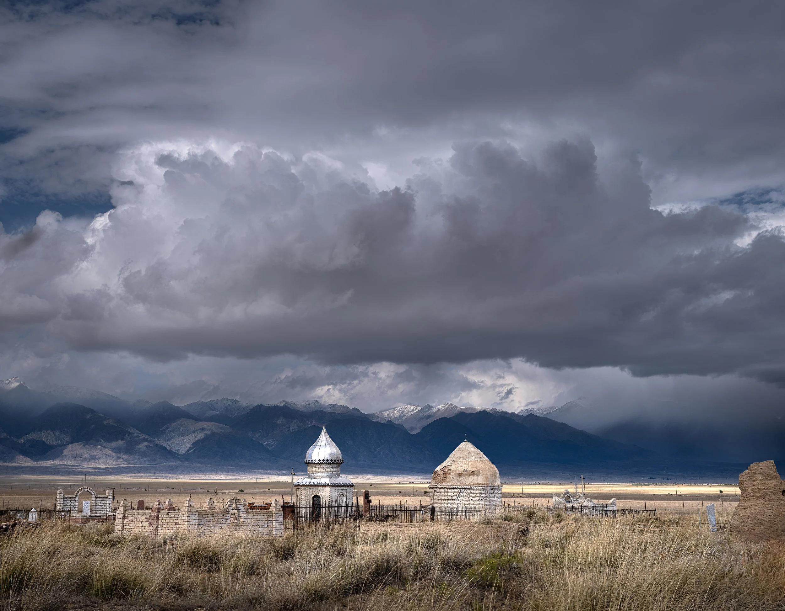 A landscape with dark, cloudy skies over mountains in the distance, with a small, white, domed building and a tower-like structure surrounded by a fence in a field of tall grass.