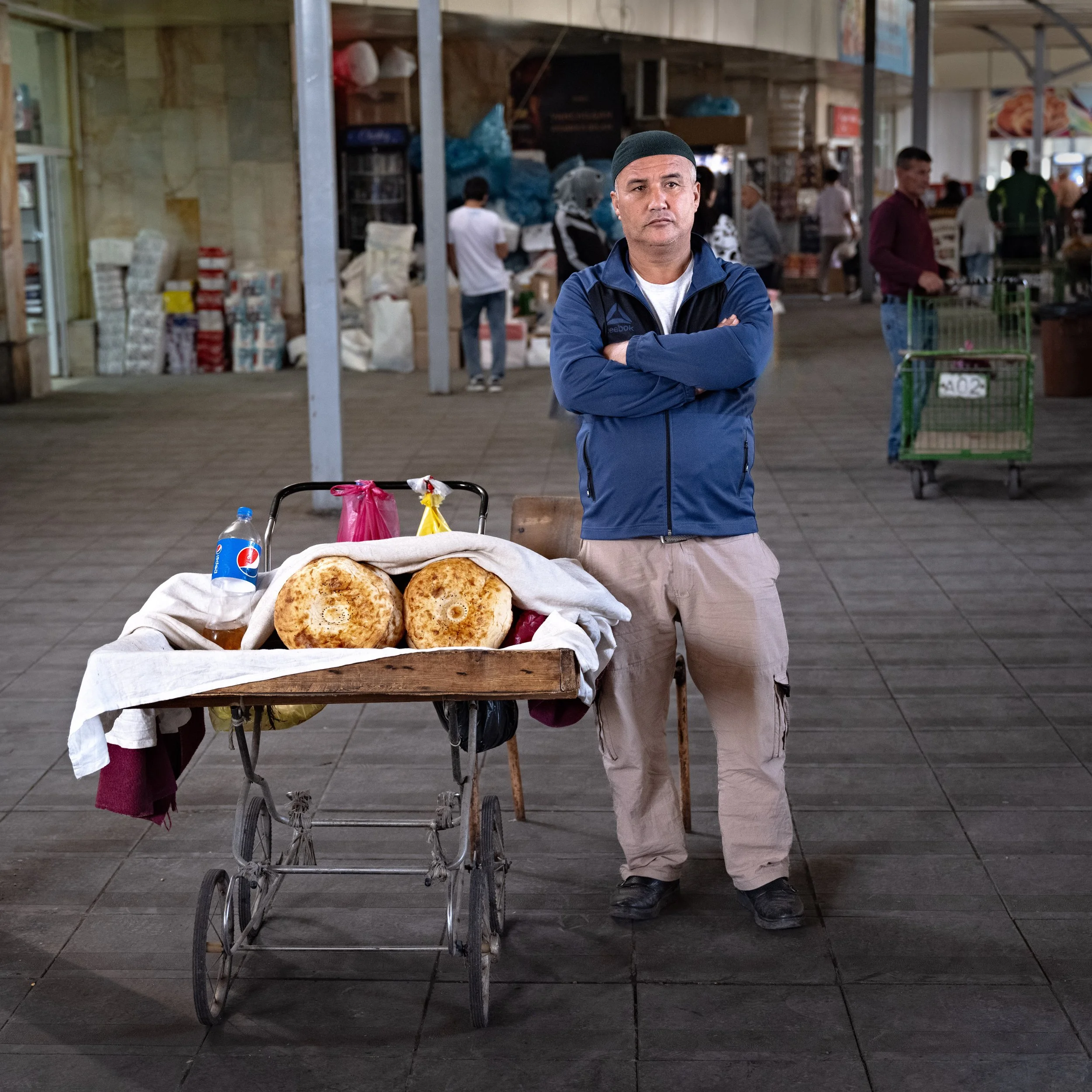 A man stands with his arms crossed behind a cart with two large round bread loaves, a bottle of Pepsi, and some plastic bags, in an indoor market with people shopping in the background.