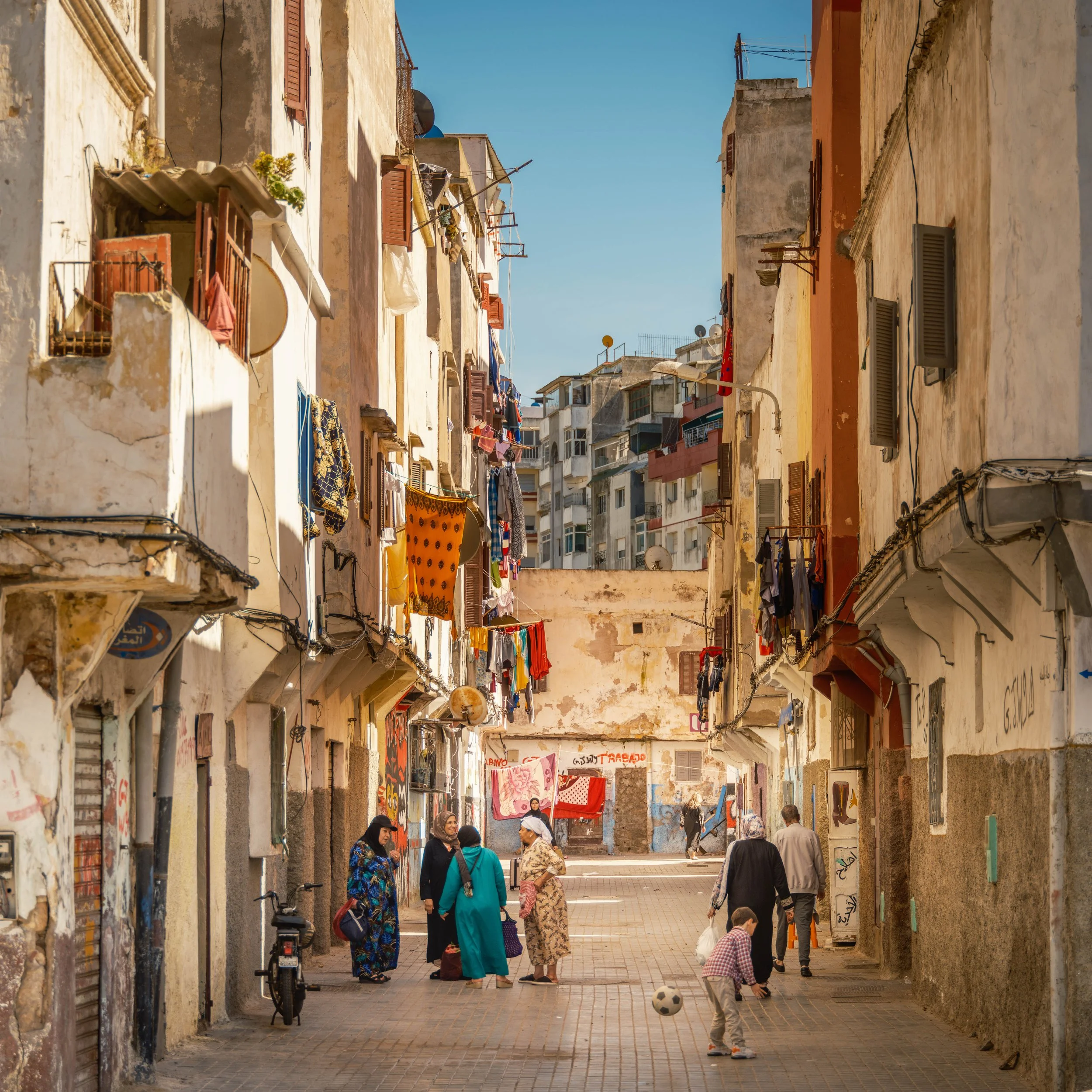 A narrow street in an urban neighborhood with old, weathered buildings, laundry hanging between the buildings, and several people walking and chatting.