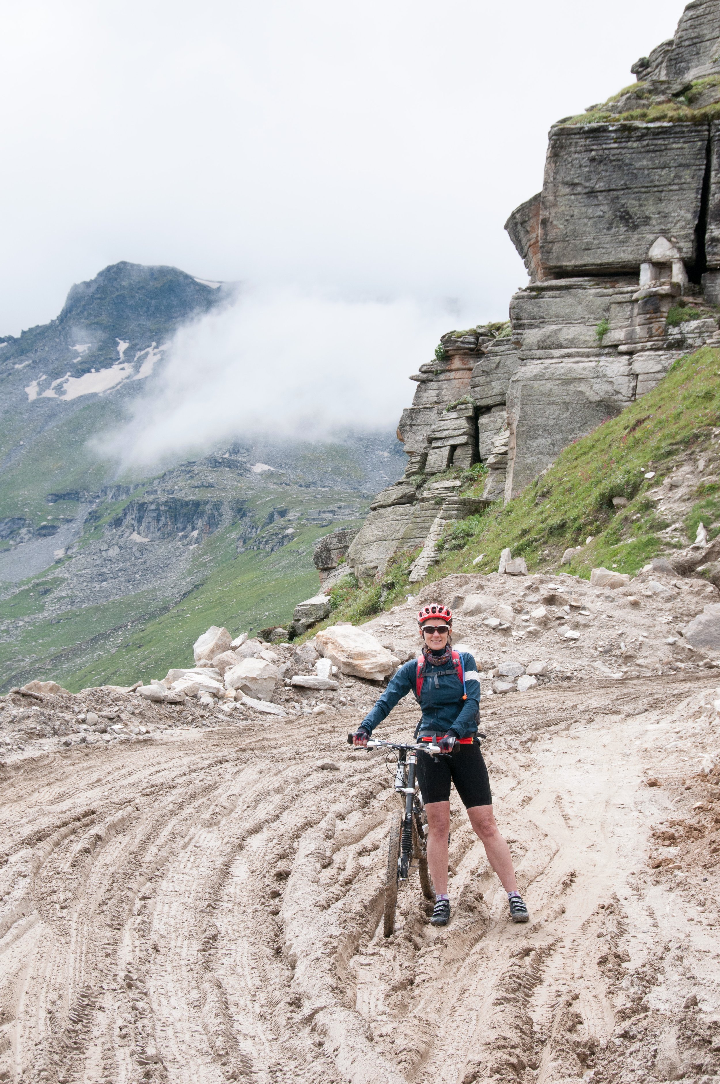 A woman with a red bike helmet, sunglasses, navy jacket, black shorts, and hiking shoes stands on a dirt trail holding a mountain bike in a mountainous landscape with clouds, rocks, and green slopes.