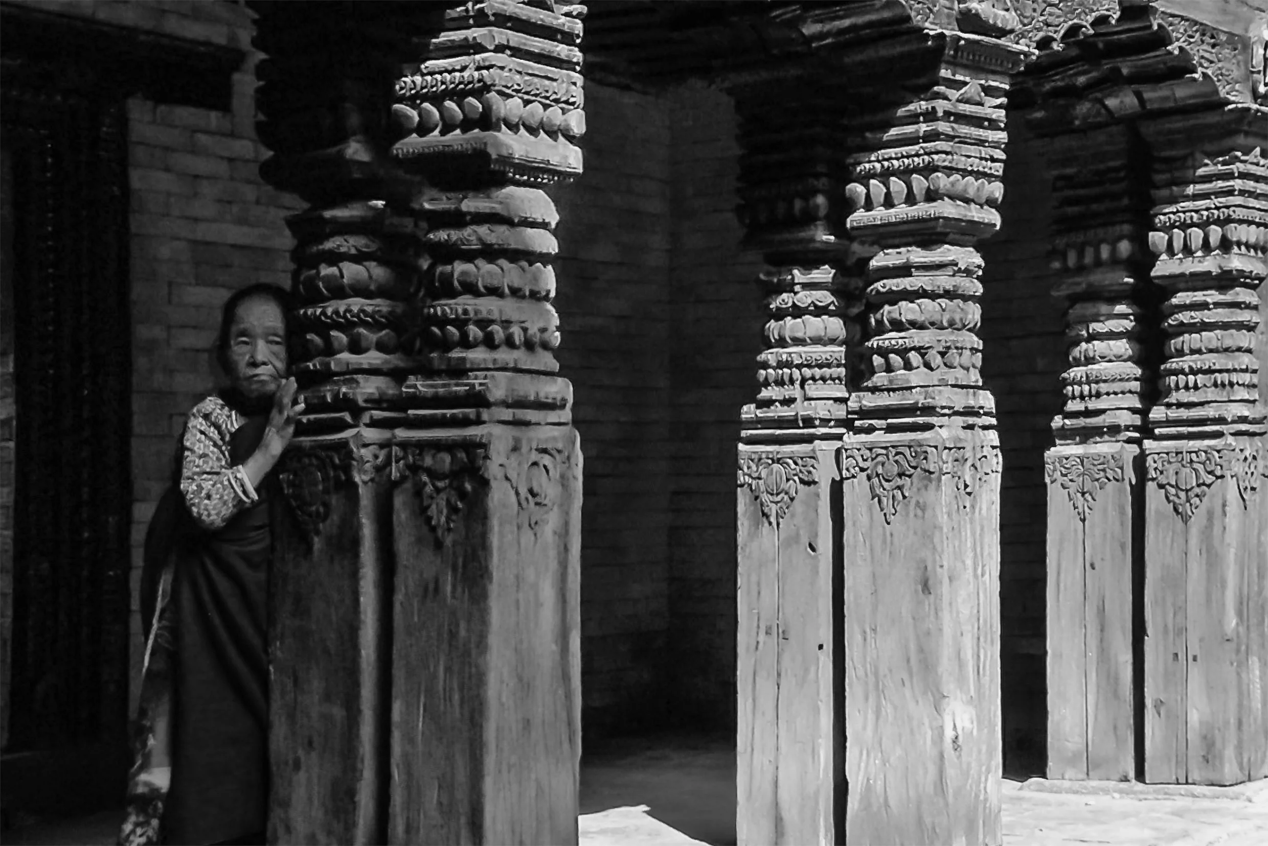 A person standing behind intricately carved wooden columns in a traditional structure, in black and white.