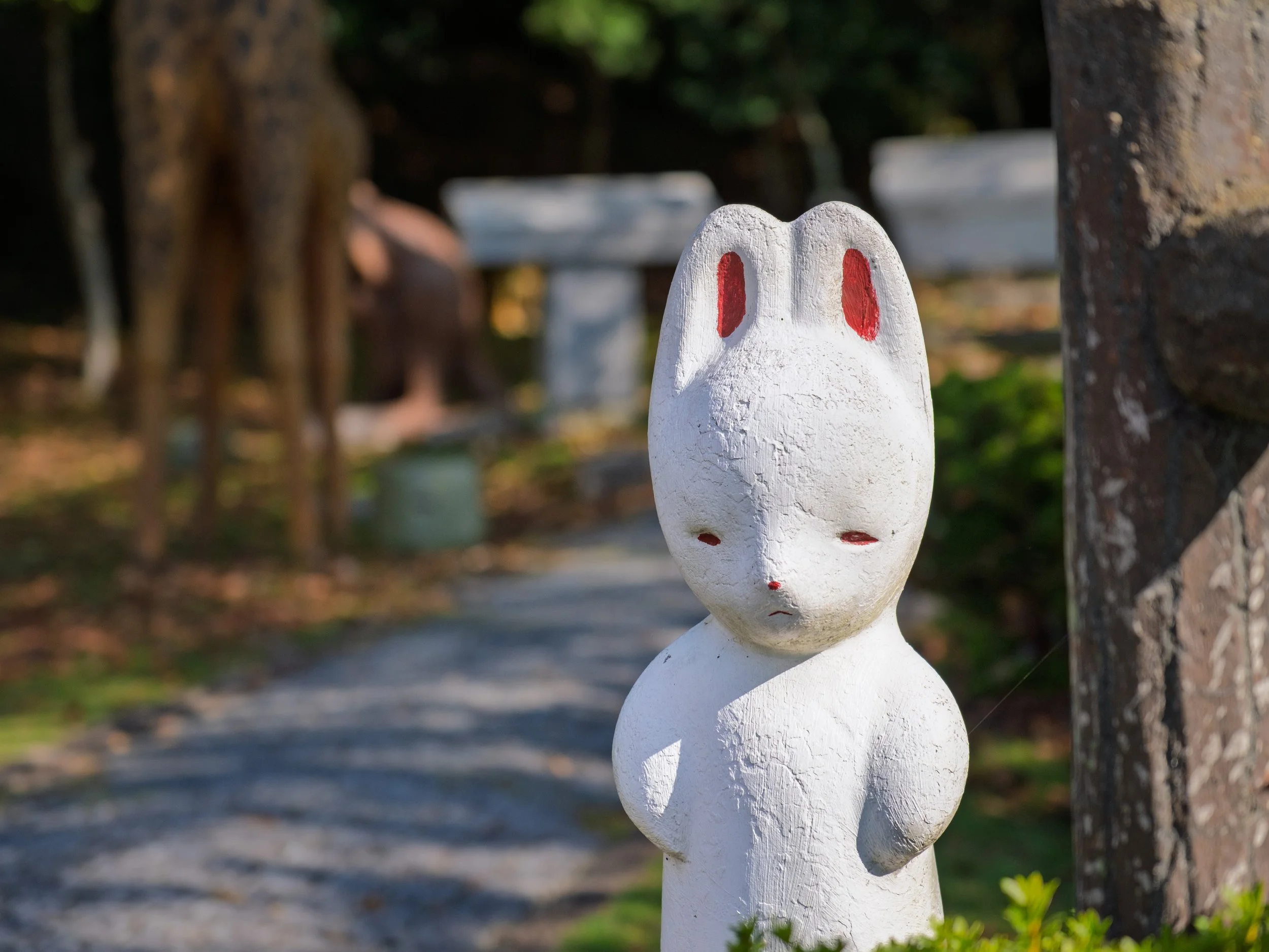 A white rabbit-shaped garden ornament with red details on its ears and small facial features, standing outdoors near a stone post with a garden path and trees in the background.