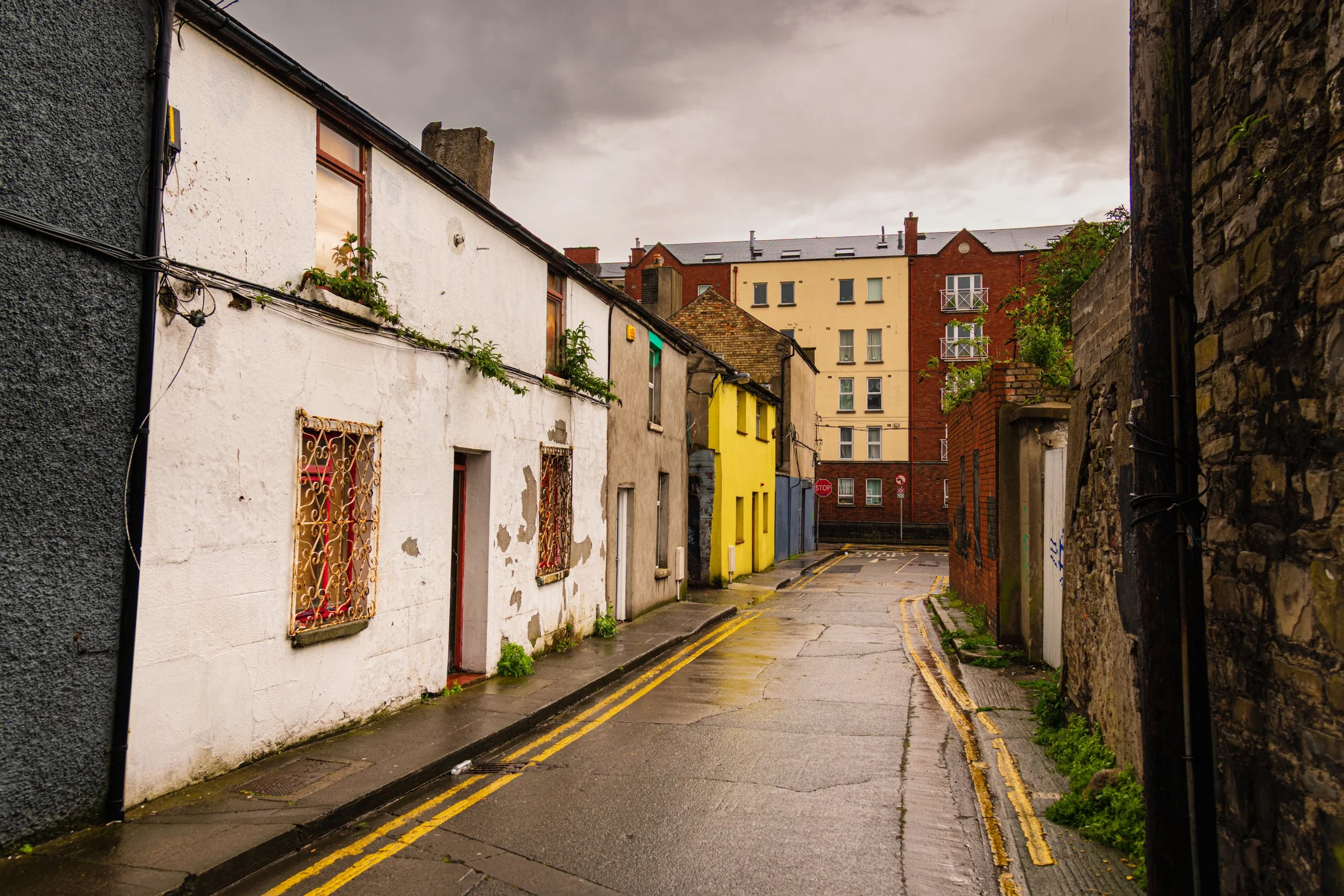 A narrow, wet city street lined with colorful buildings and stone walls, with cloudy skies overhead.