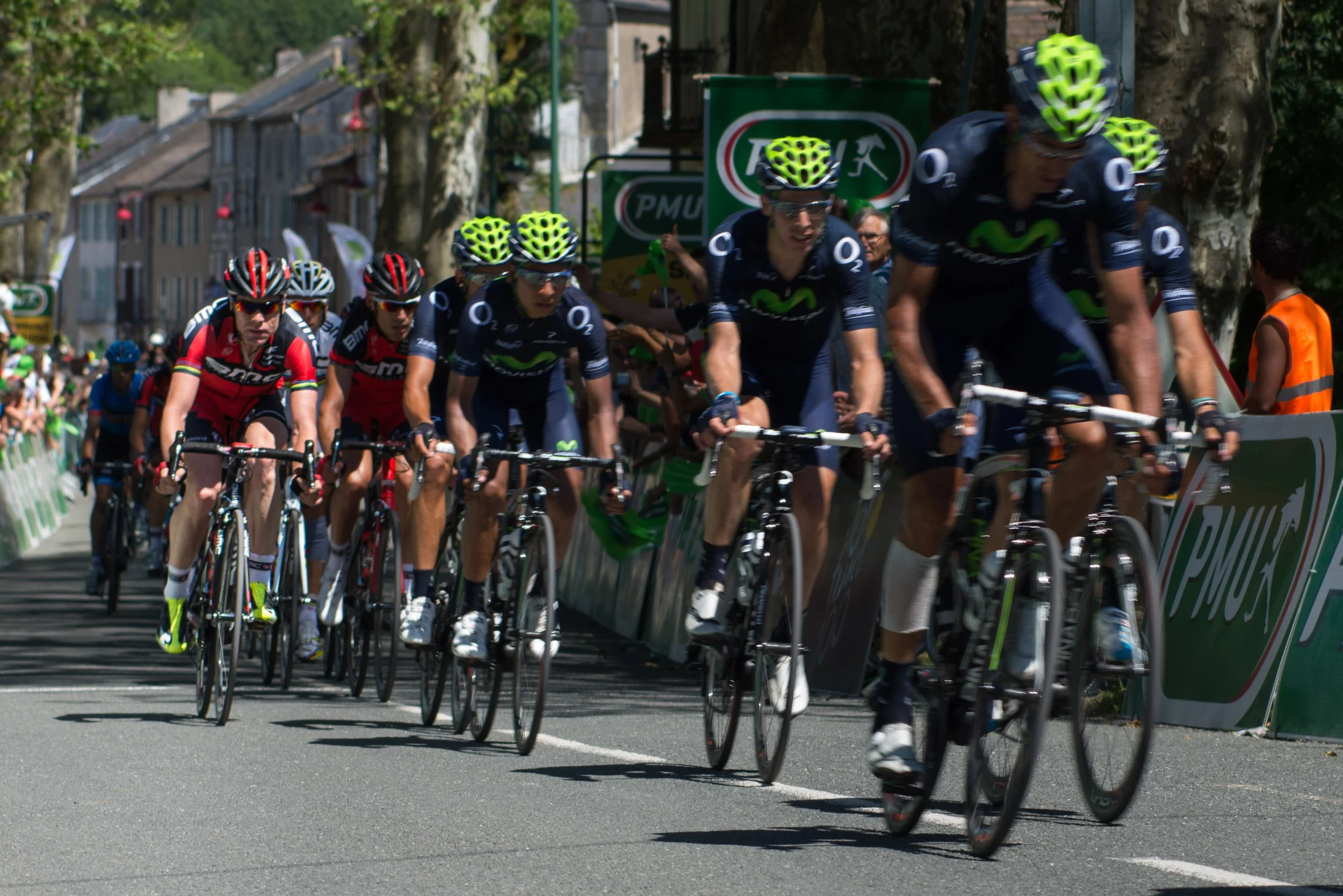 A group of cyclists racing on a city street during daytime, wearing helmets and team jerseys, with spectators watching from the sides.