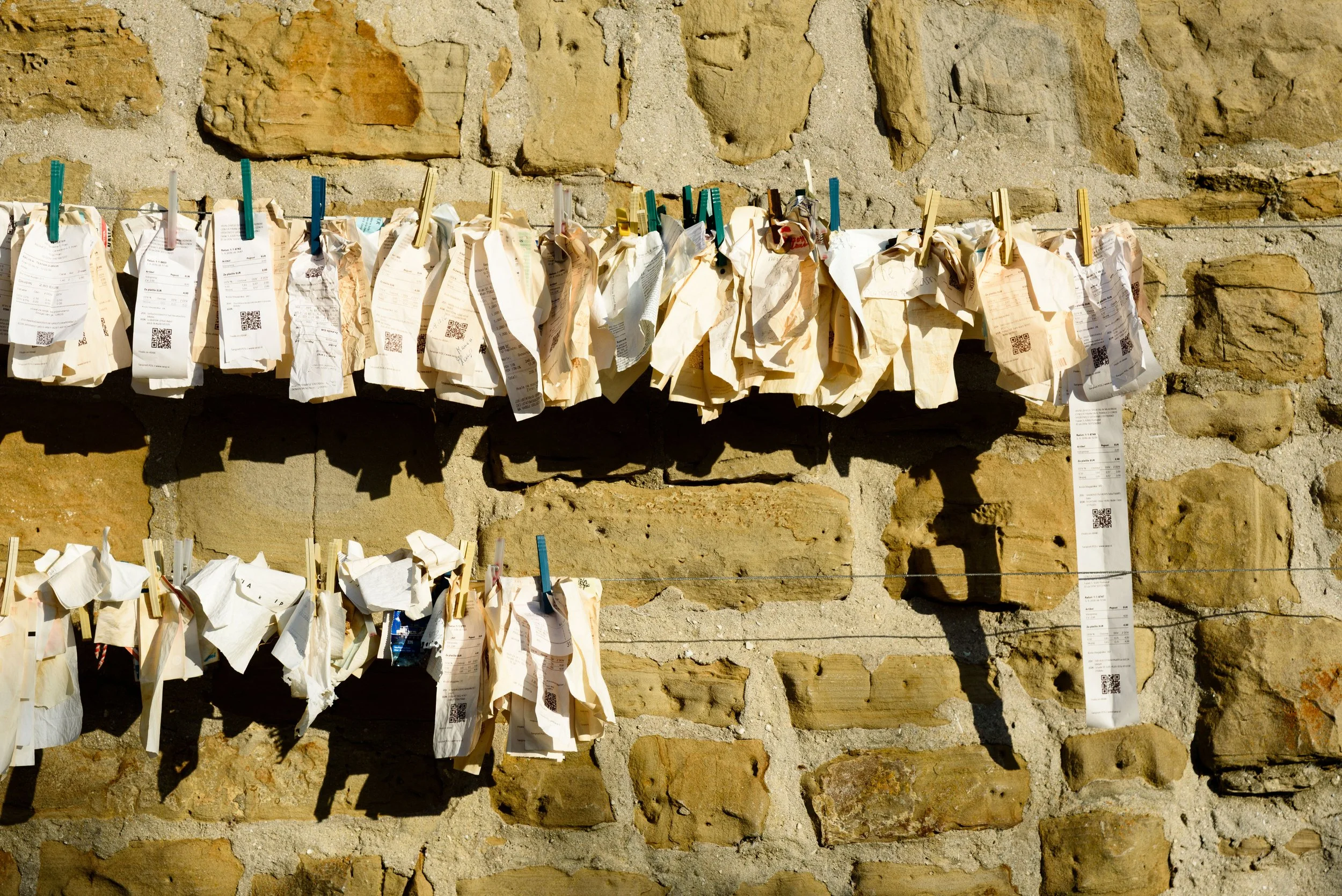 Multiple crumpled paper receipts hanging on clotheslines with clothespins against a stone wall.