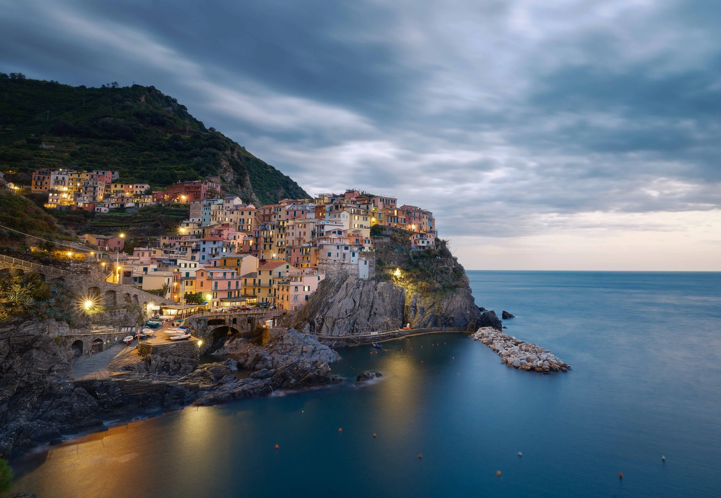Colorful hillside town with buildings illuminated by lights, overlooking a calm ocean under a stormy sky at dusk.