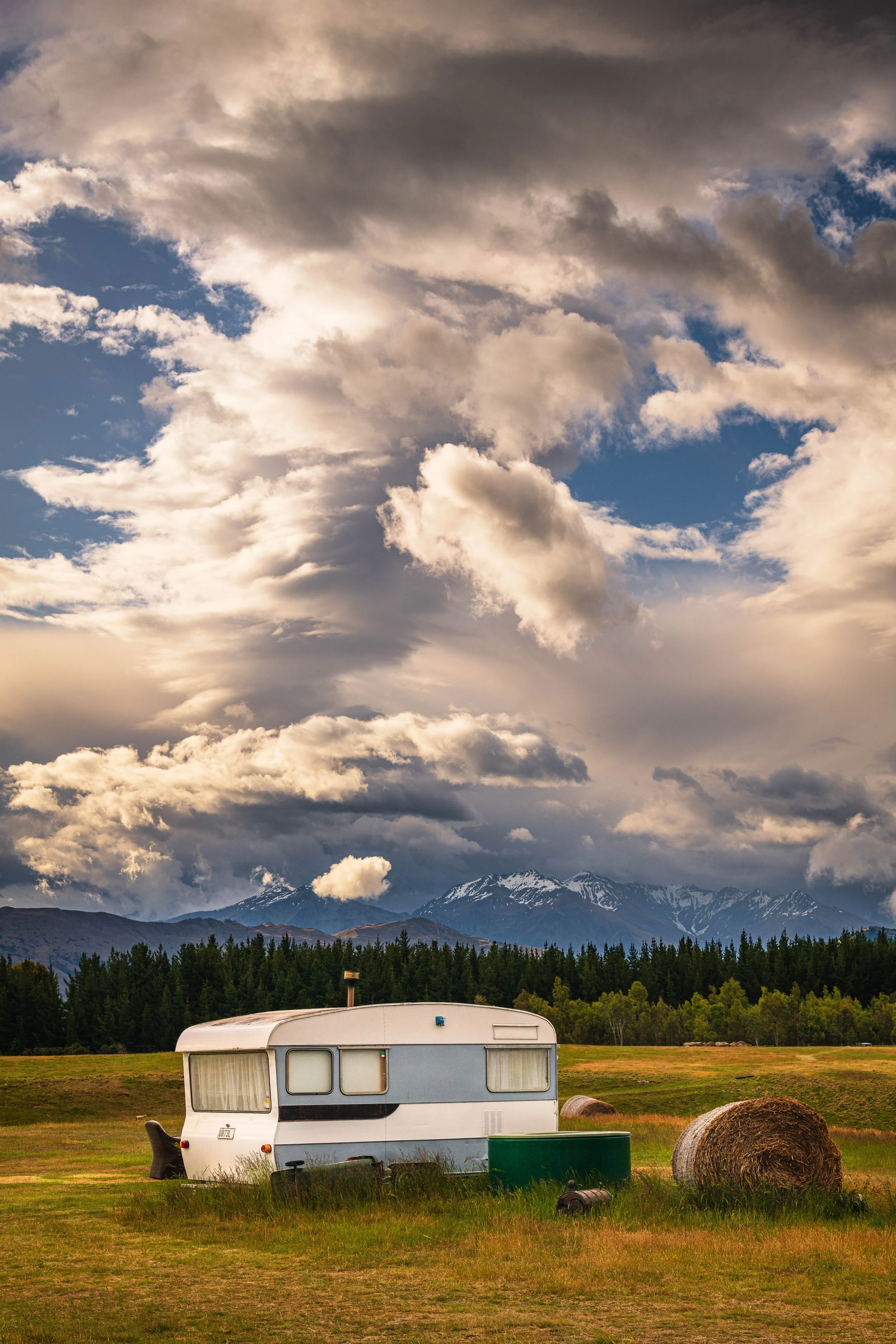 An empty travel trailer parked in a grassy field with hay bales, with evergreen trees, snow-capped mountains, and a cloudy sky in the background.