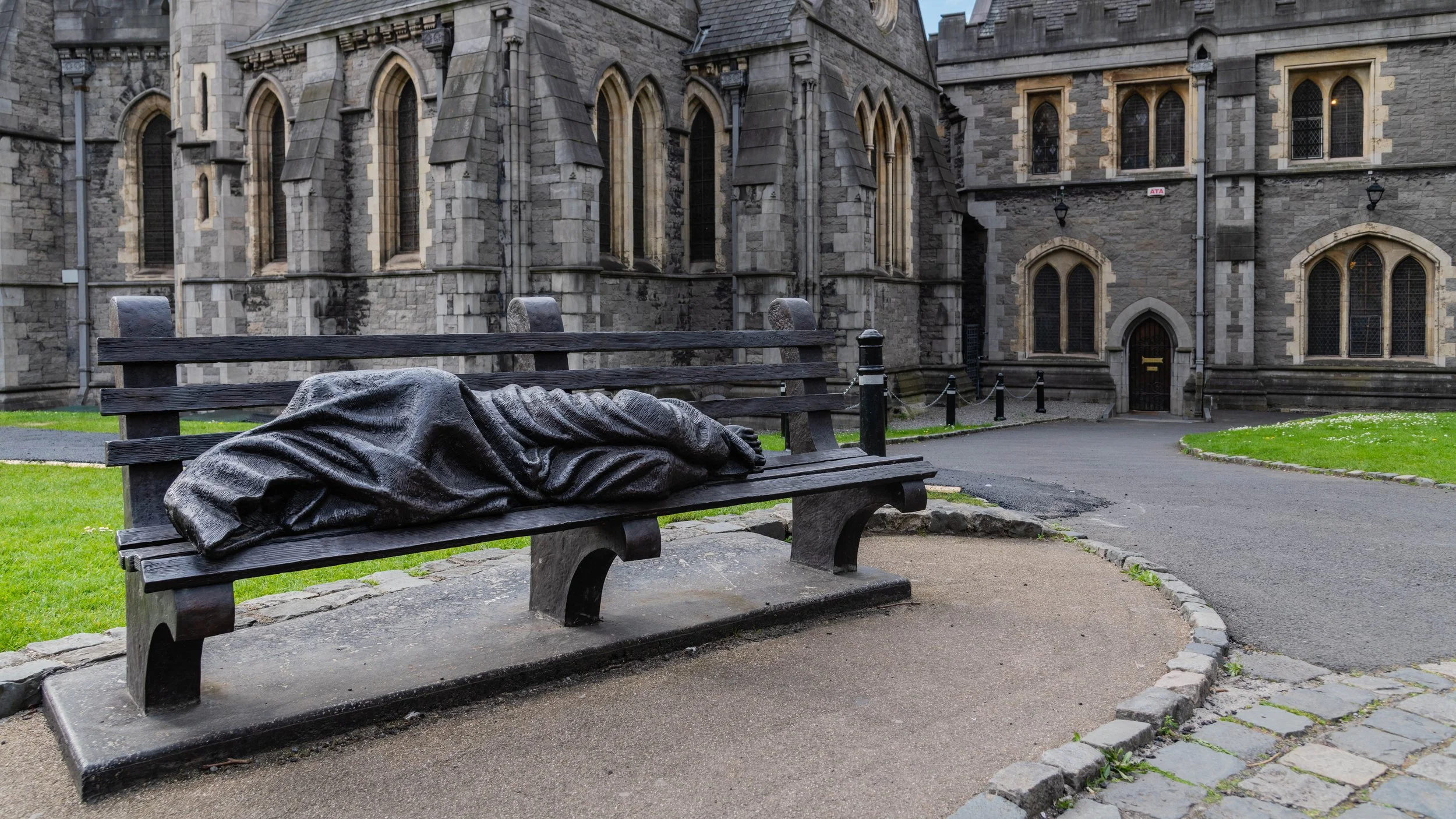 A bronze sculpture of a man lying on a park bench in front of a historic stone church.