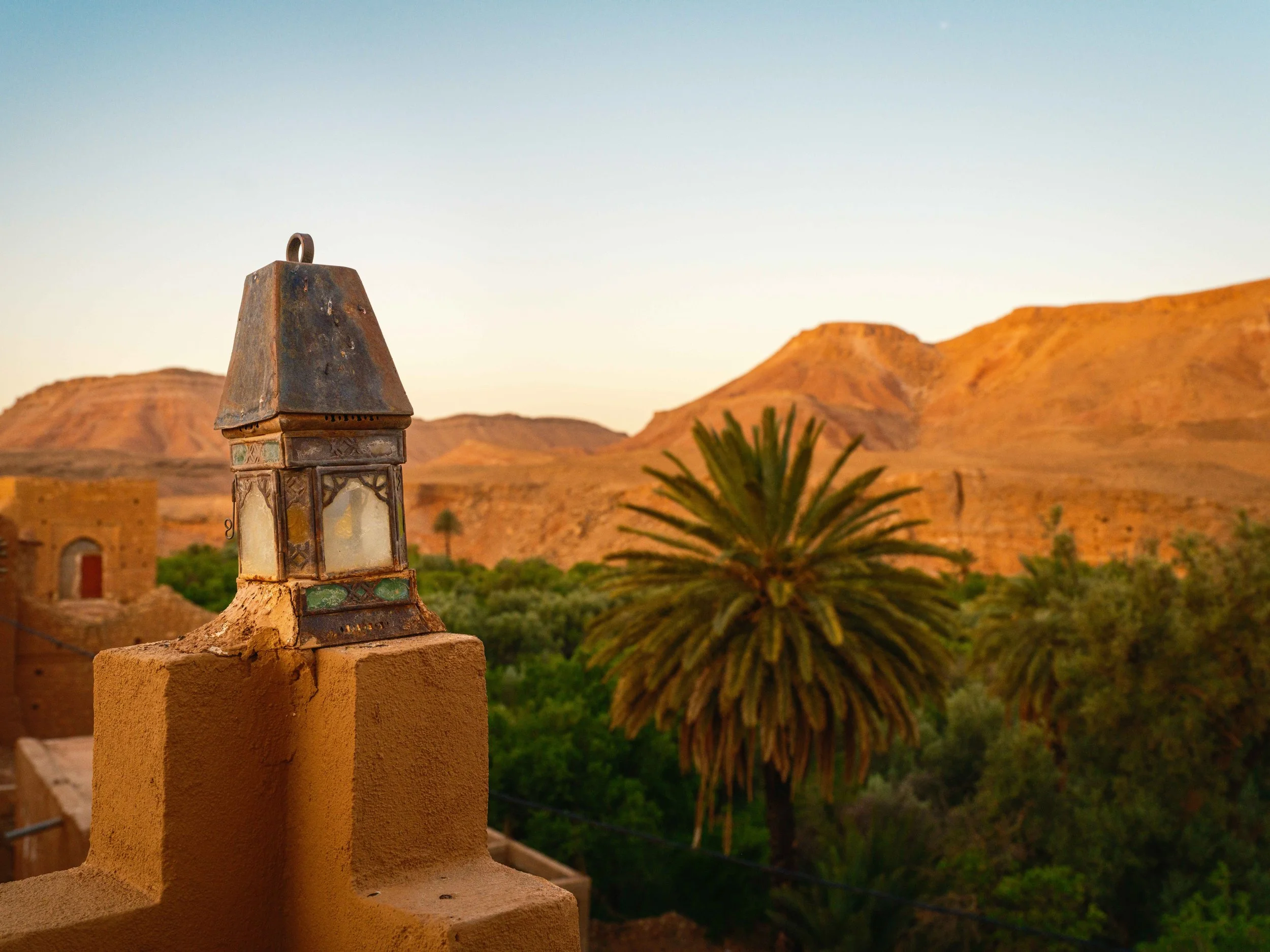 A rustic lantern on a rooftop with desert hills and palm trees in the distance during sunset.