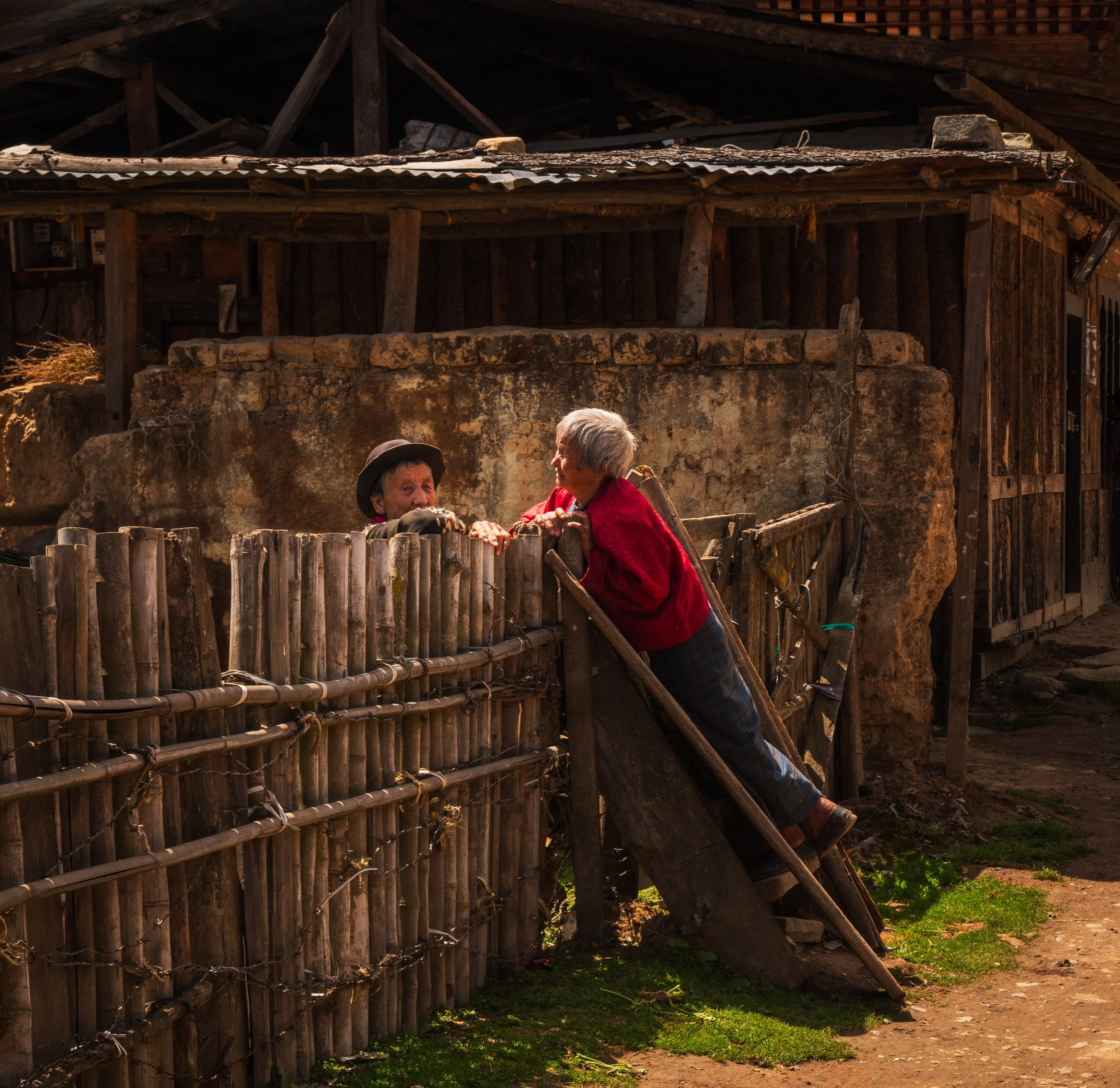 Two elderly women leaning over a bamboo fence and conversing outside a rustic wooden building during daytime.