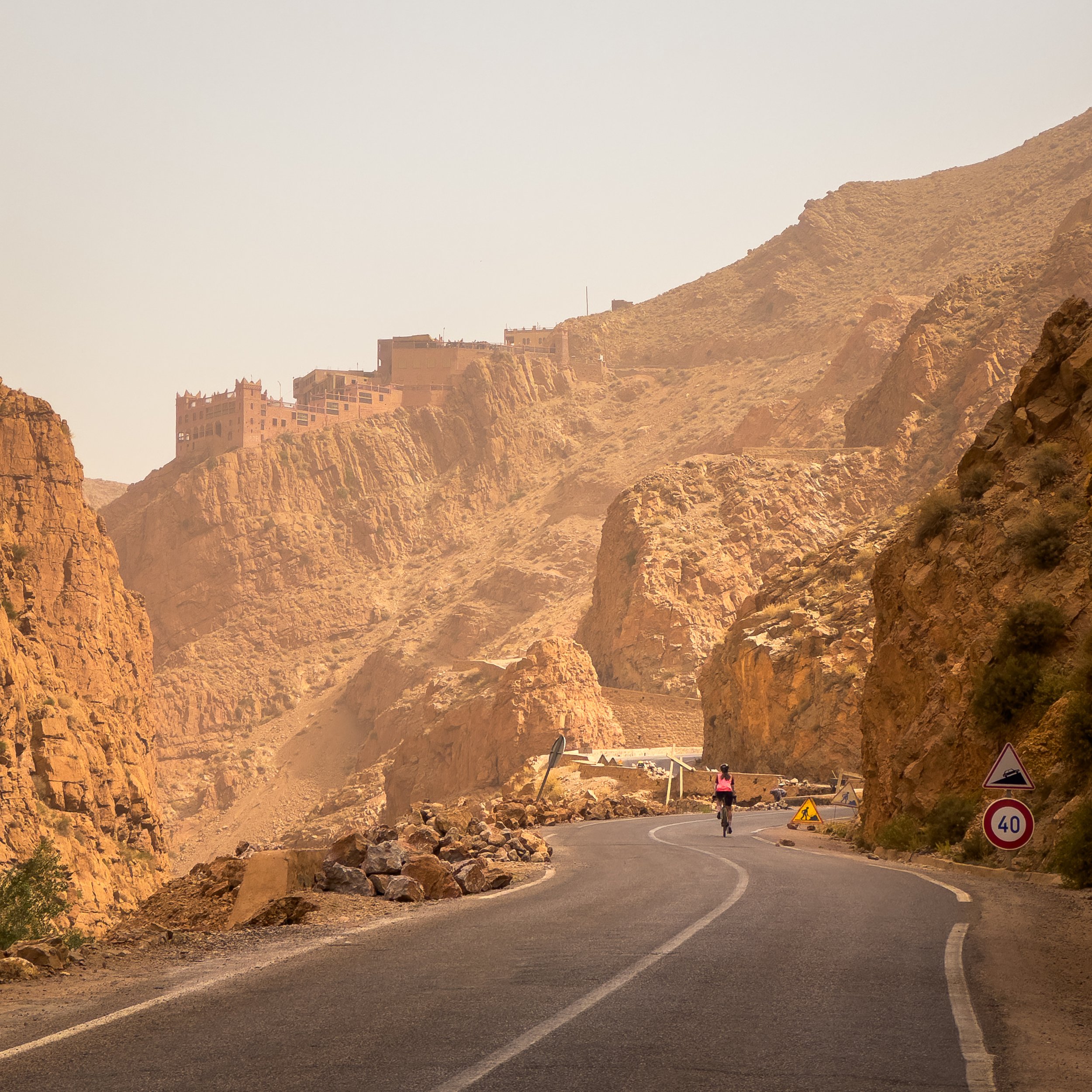 A winding mountain road with a cyclist riding away from the camera, surrounded by rocky cliffs and a village on a hilltop.