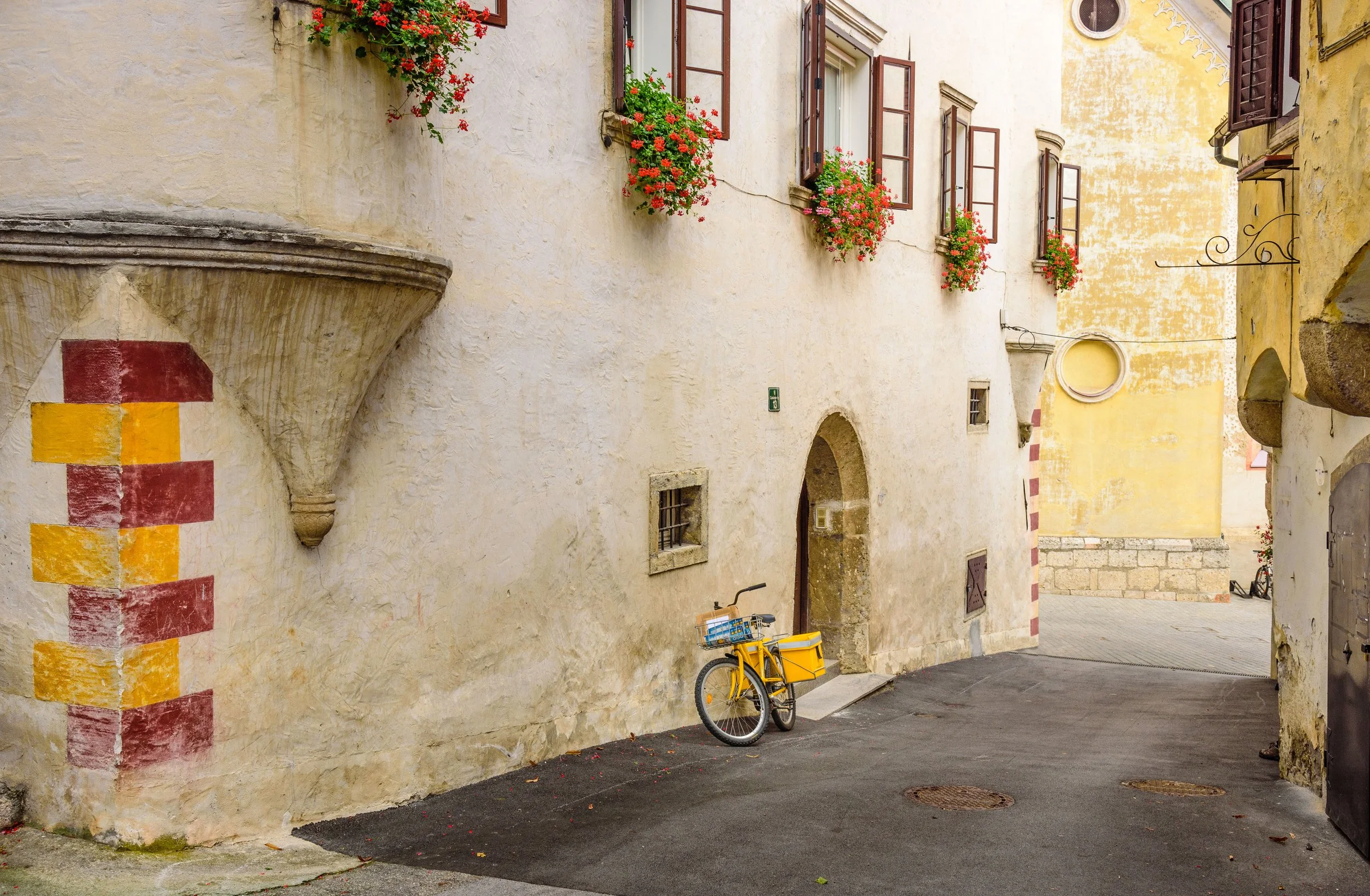 A yellow bicycle with a basket parked against a textured, cream-colored wall in an old European alleyway with flower boxes on windows and a mural on the wall.