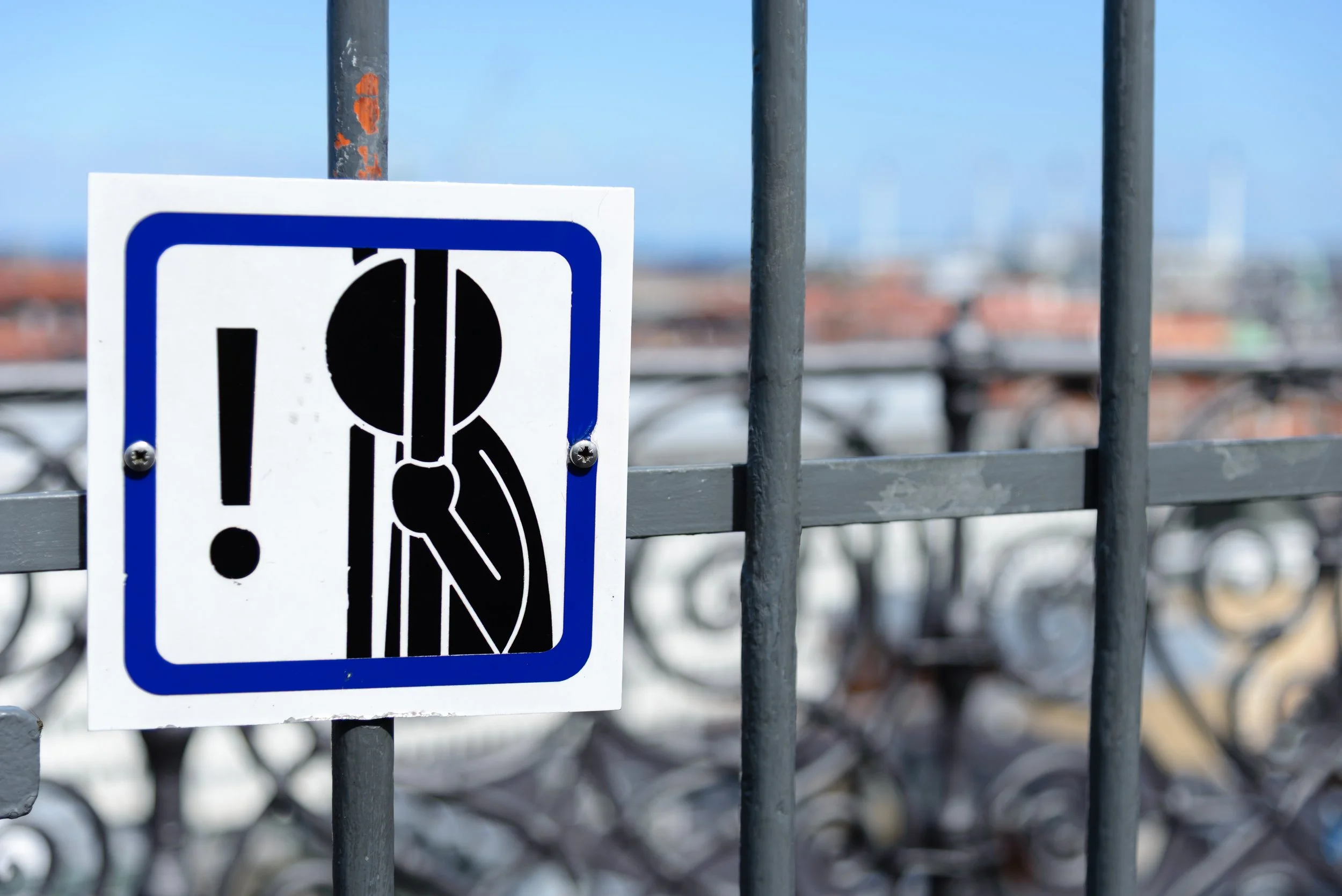 Close-up of a warning sign on a metal fence with blurred bicycles and sky in the background. The sign shows a person looking through a safety device and an exclamation mark.