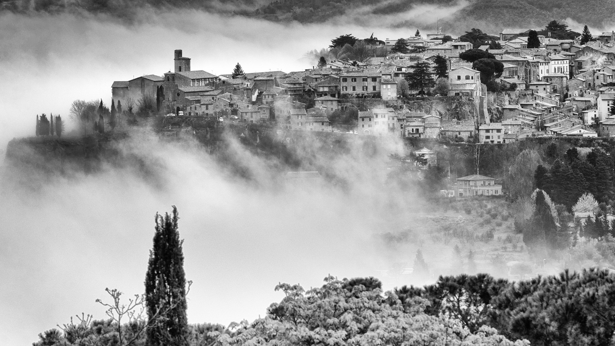 Black and white photo of a hillside village surrounded by fog, with buildings, trees, and a church visible.