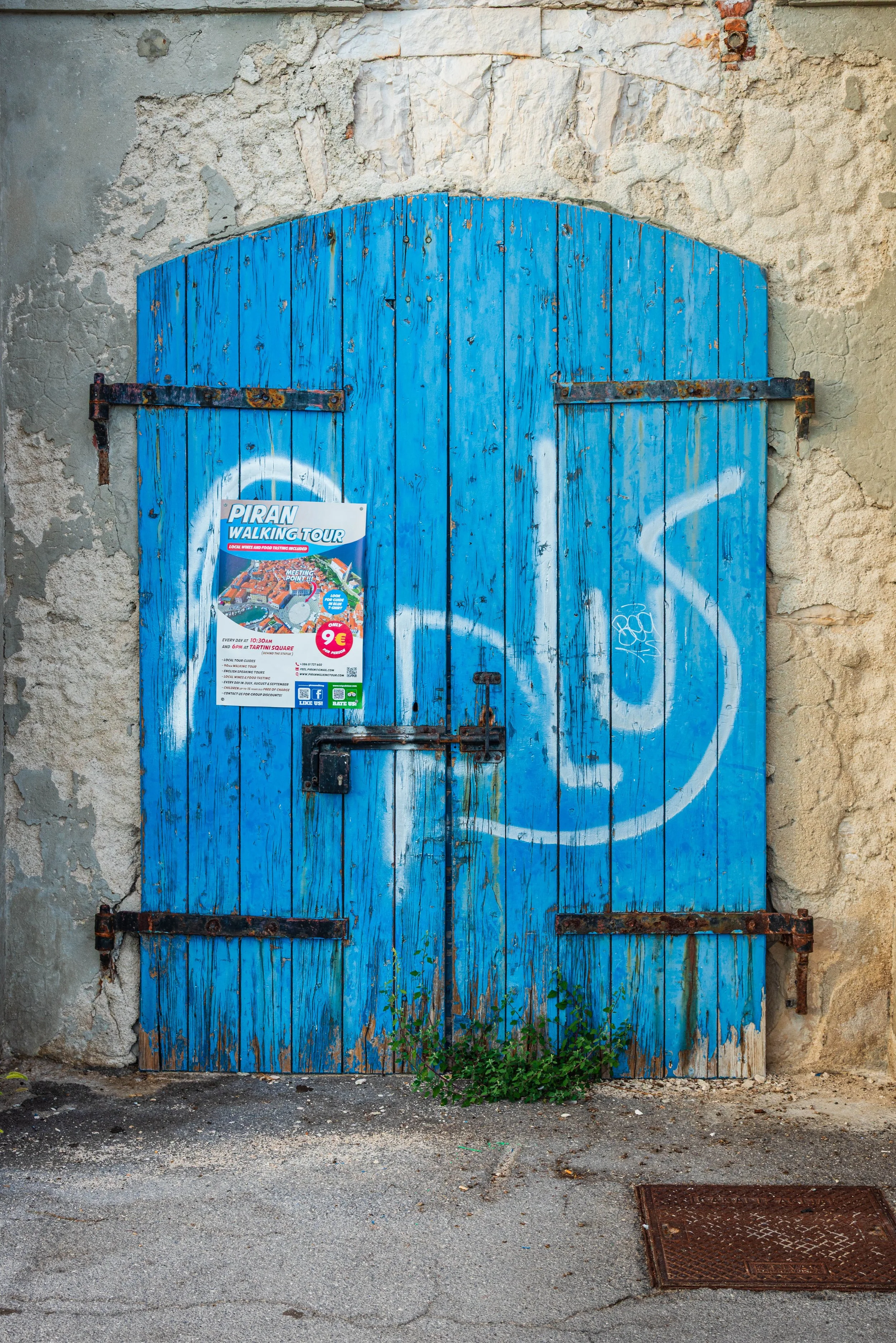 Blue wooden door with graffiti, rusted hinges, a padlock, and a poster advertising a walking tour. The door is set in a weathered stone wall with plants growing at its base.