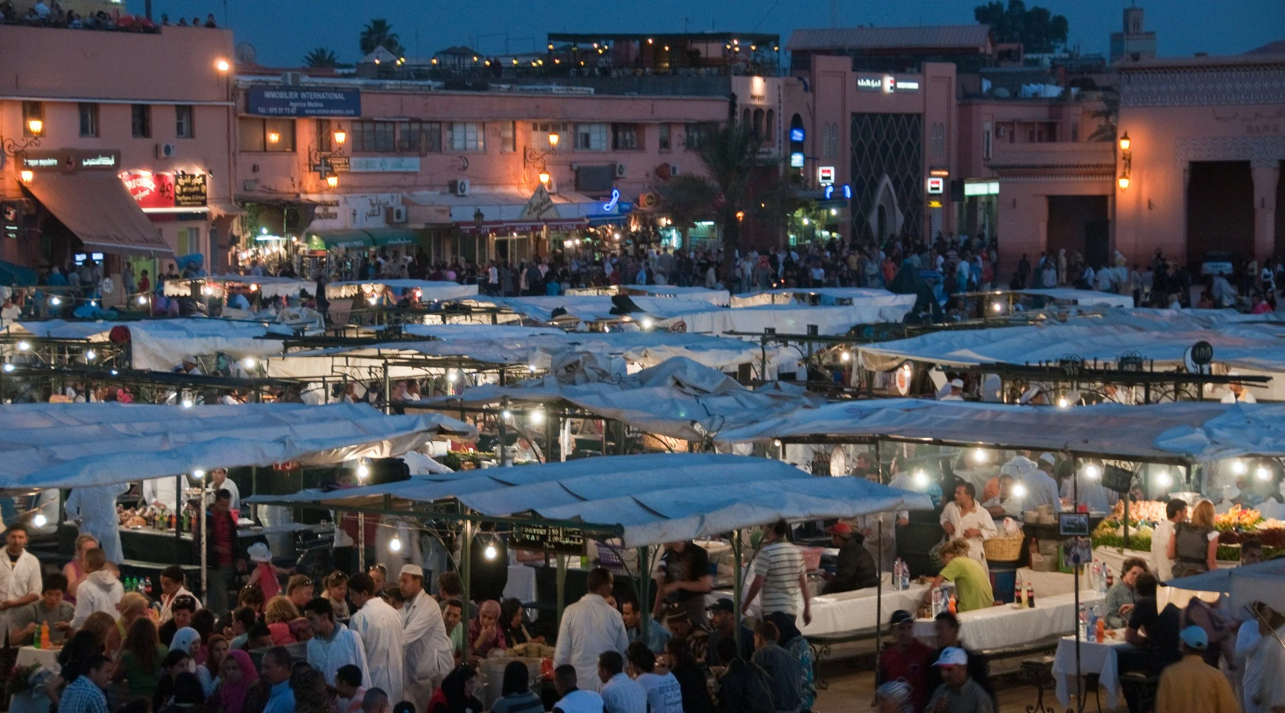 Night market scene with crowded outdoor stalls, many people shopping and dining, surrounding buildings illuminated in pink and orange, some with signs and lights, under a darkening sky.
