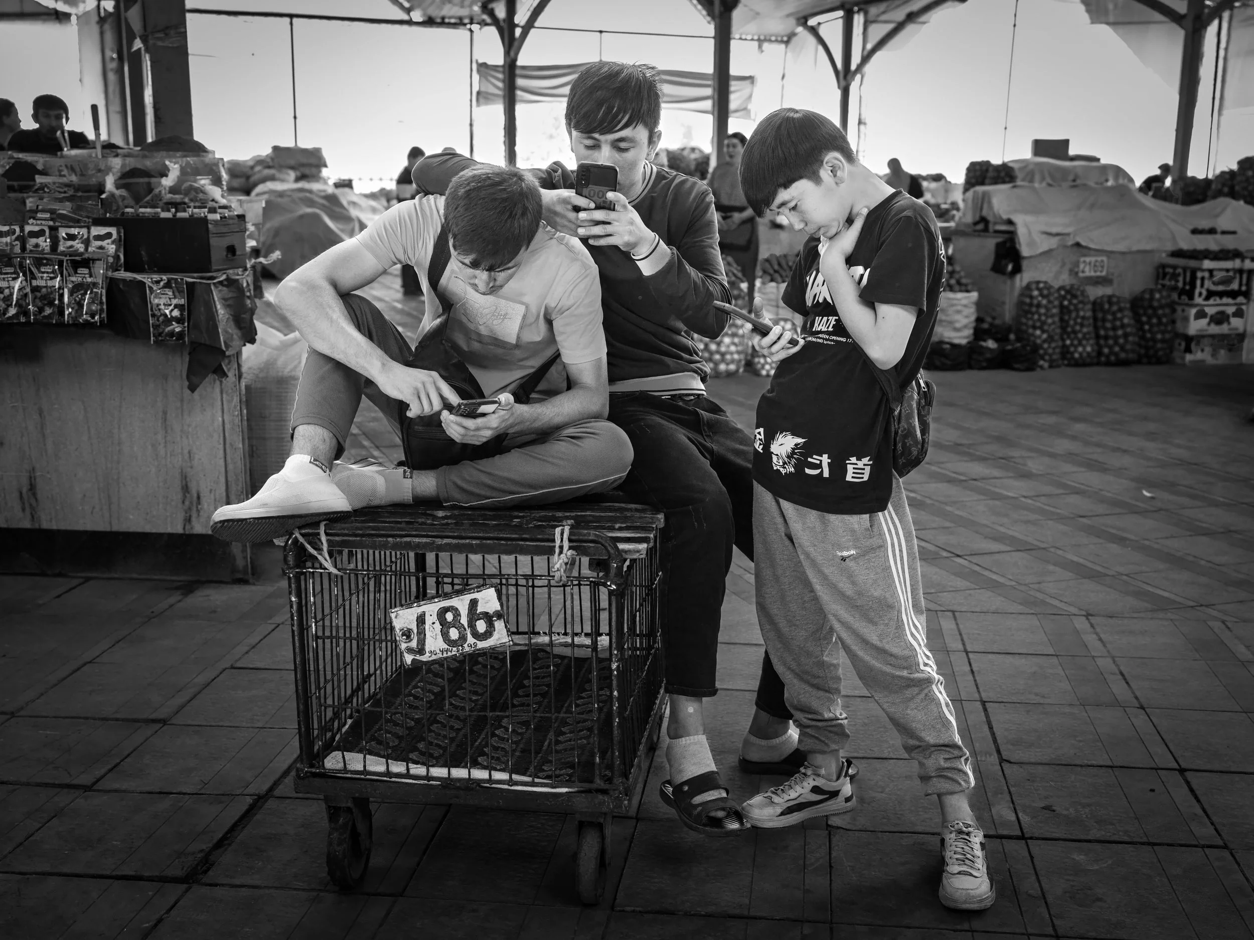Three young males engaging with their smartphones in a market setting, with stalls and merchandise around them in a tented area.