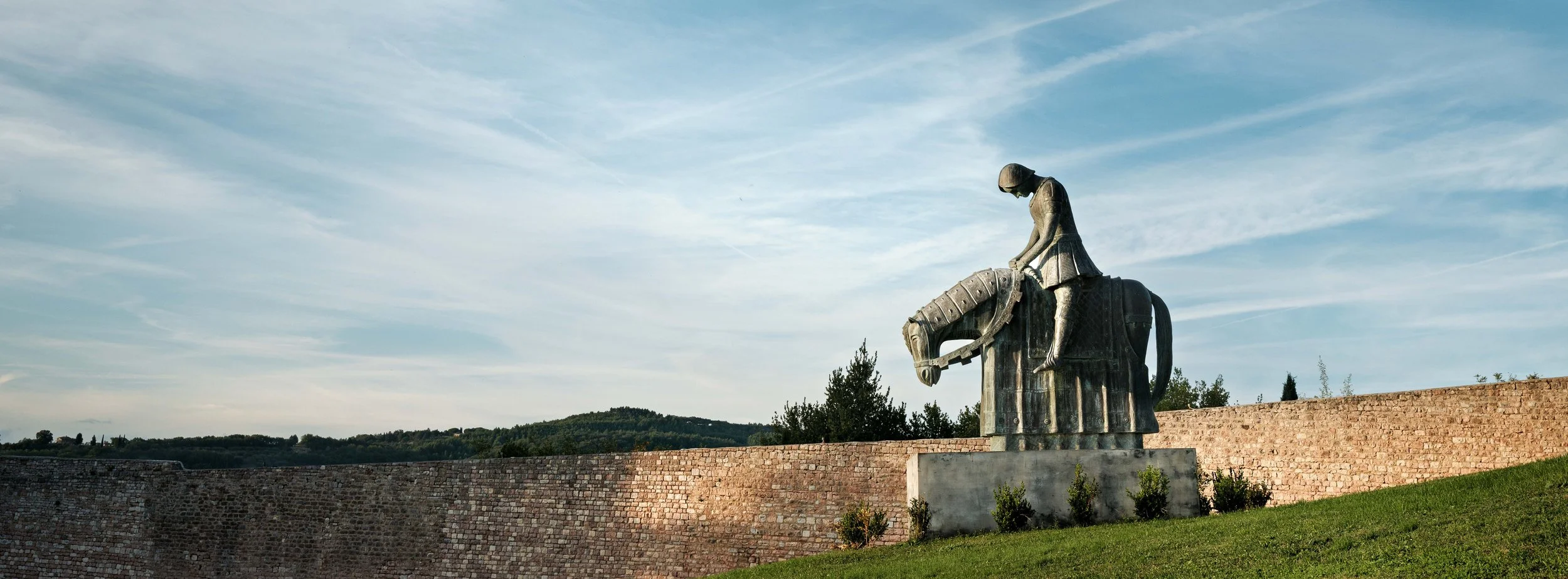 Statue of a woman riding a horse, located outdoors against a backdrop of a brick wall and a partly cloudy sky.