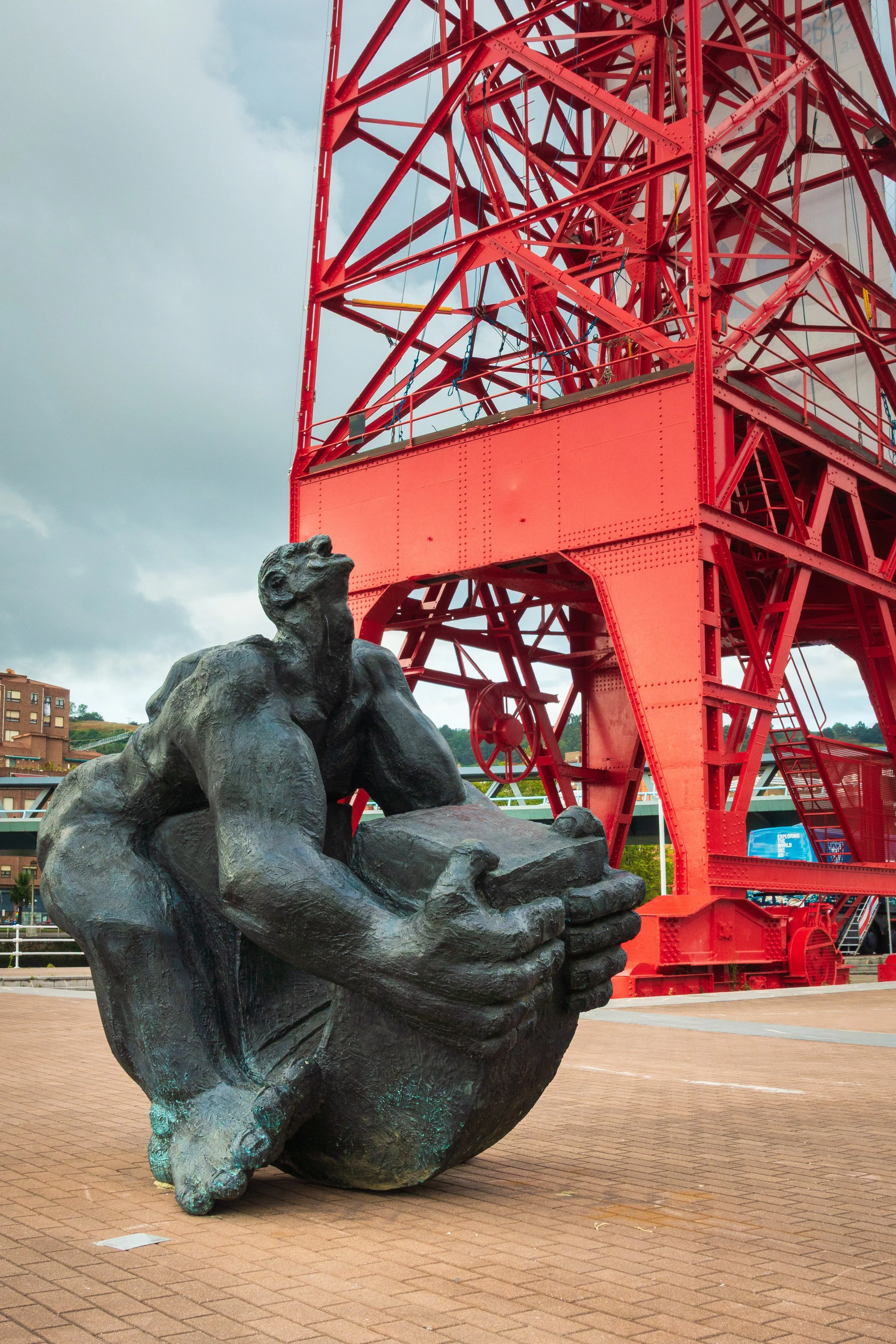 A sculpture of two large hands holding a rock, with a statue of a person sitting on a large gear in front, next to a tall red metal structure, possibly a historical industrial site or landmark.