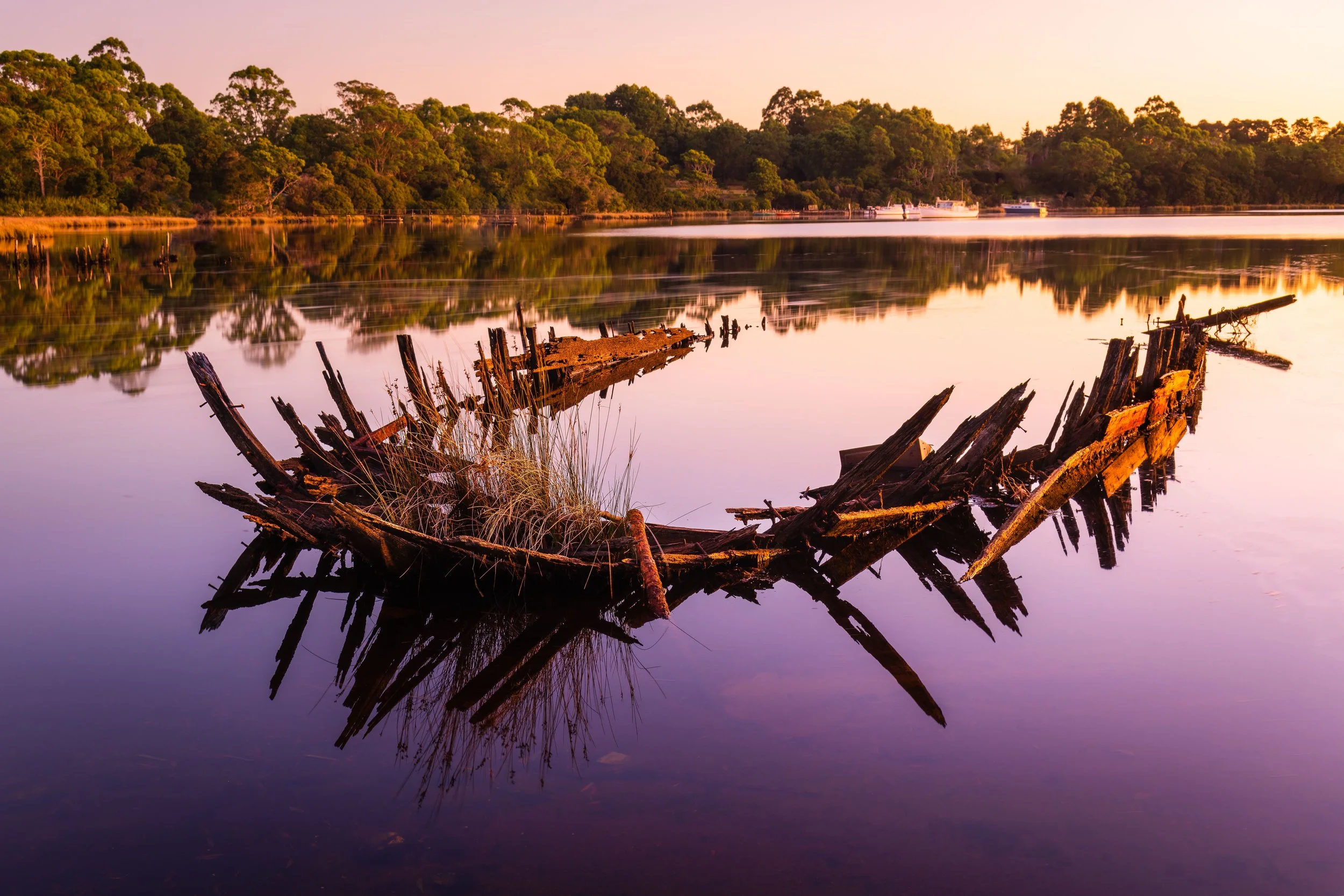 Sunset over a calm river with a partially submerged, broken wooden dock in the foreground and a dense green wooded shoreline in the background.