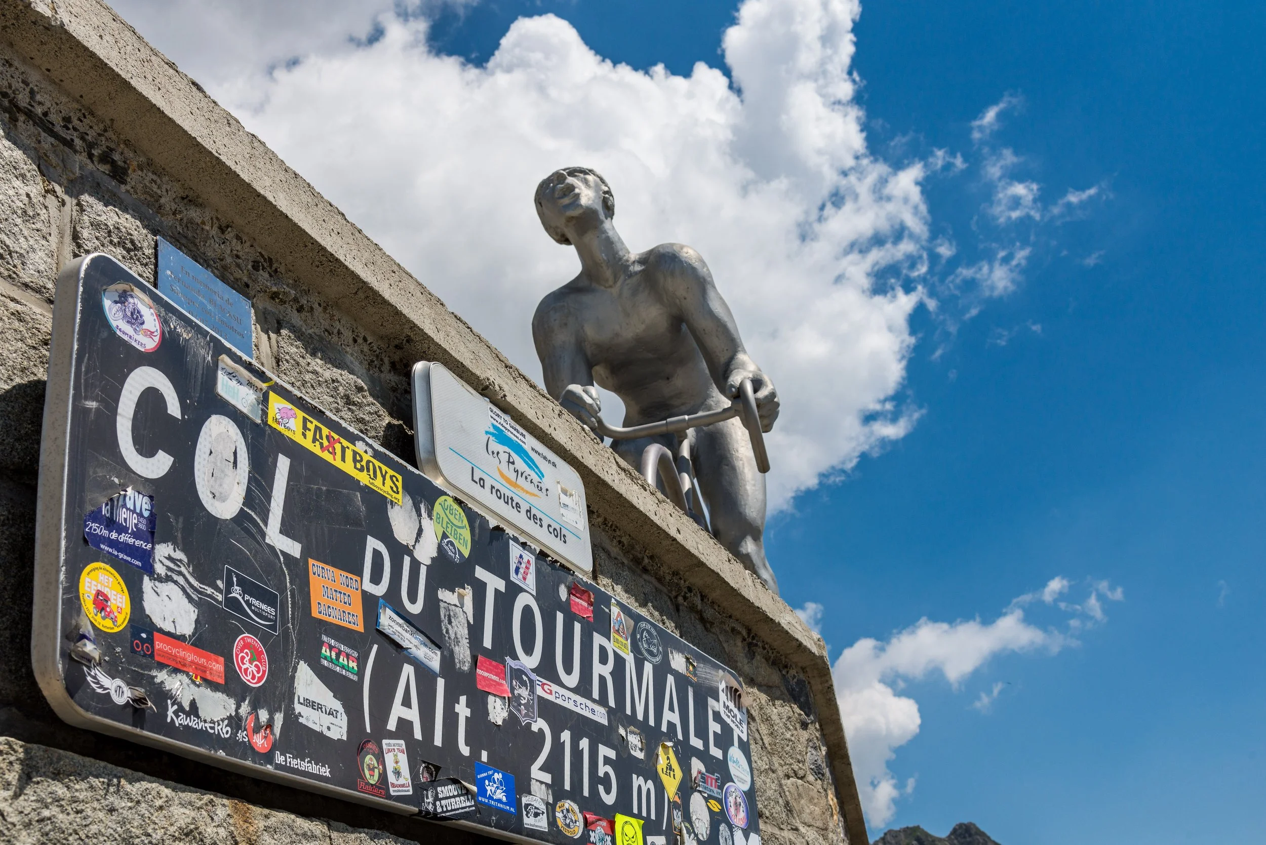 Street sign with numerous stickers reading 'Col du Tourmalet, Alt. 2115 m' beneath a statue of a cyclist on a stone wall, against a blue sky with clouds.