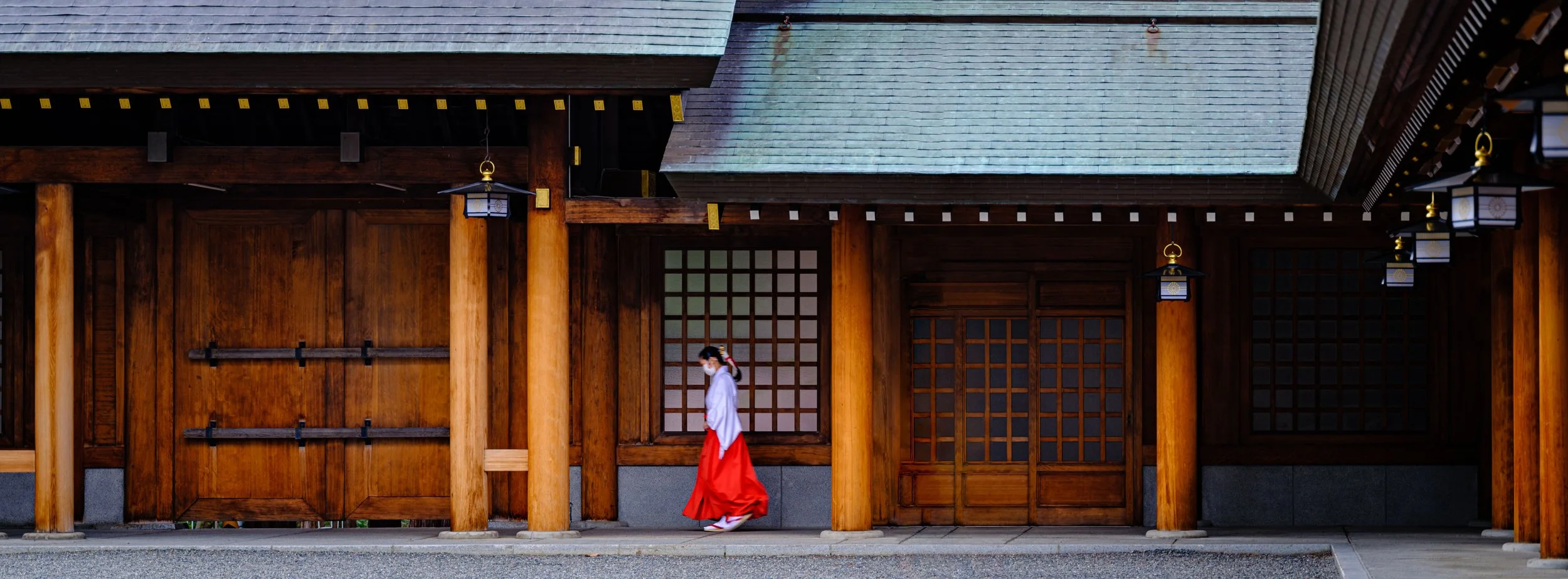 A woman dressed in traditional Japanese attire walking past a wooden building with shoji screens and hanging lanterns.