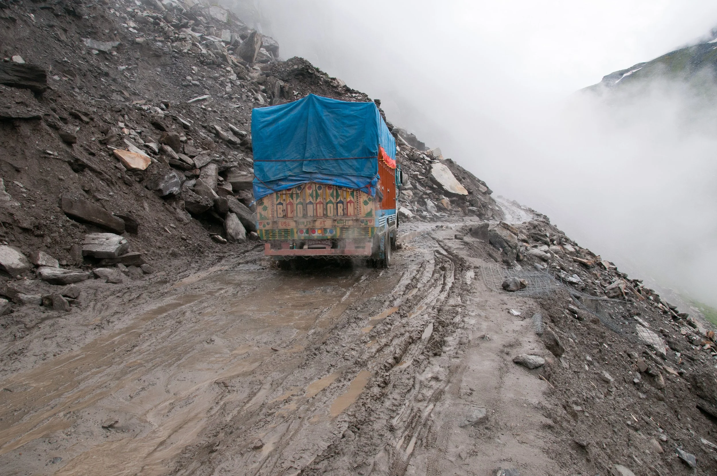 A colorful truck with a blue tarp cover driving on a muddy mountain road surrounded by rocky terrain and fog.