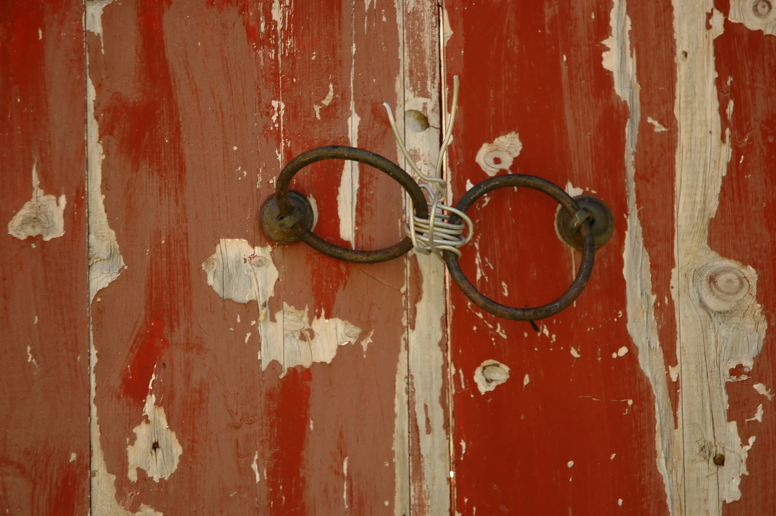 Rusty metal barn door with peeling red paint, tied together with wire and a large metal hook.